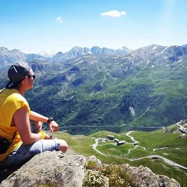 A person sitting on a rock looking at a scenic mountain landscape with green hills, winding roads, and distant snow-capped peaks.