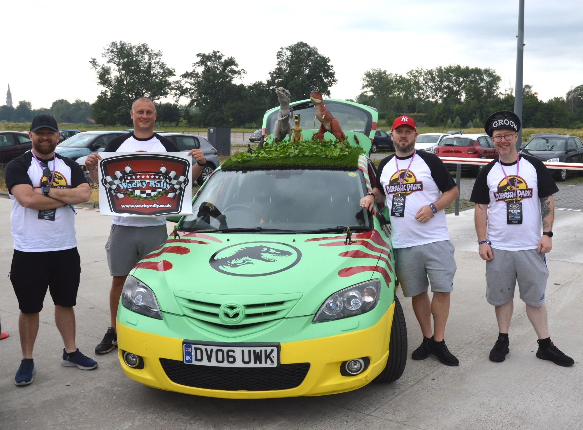 Group of five men standing around a decorated car at a rally event. The car has a green and yellow dinosaur-themed design with Jurassic Park logos and toy dinosaurs on top. One man is holding a 'Wacky Rally' sign, and they are all wearing event T-shi