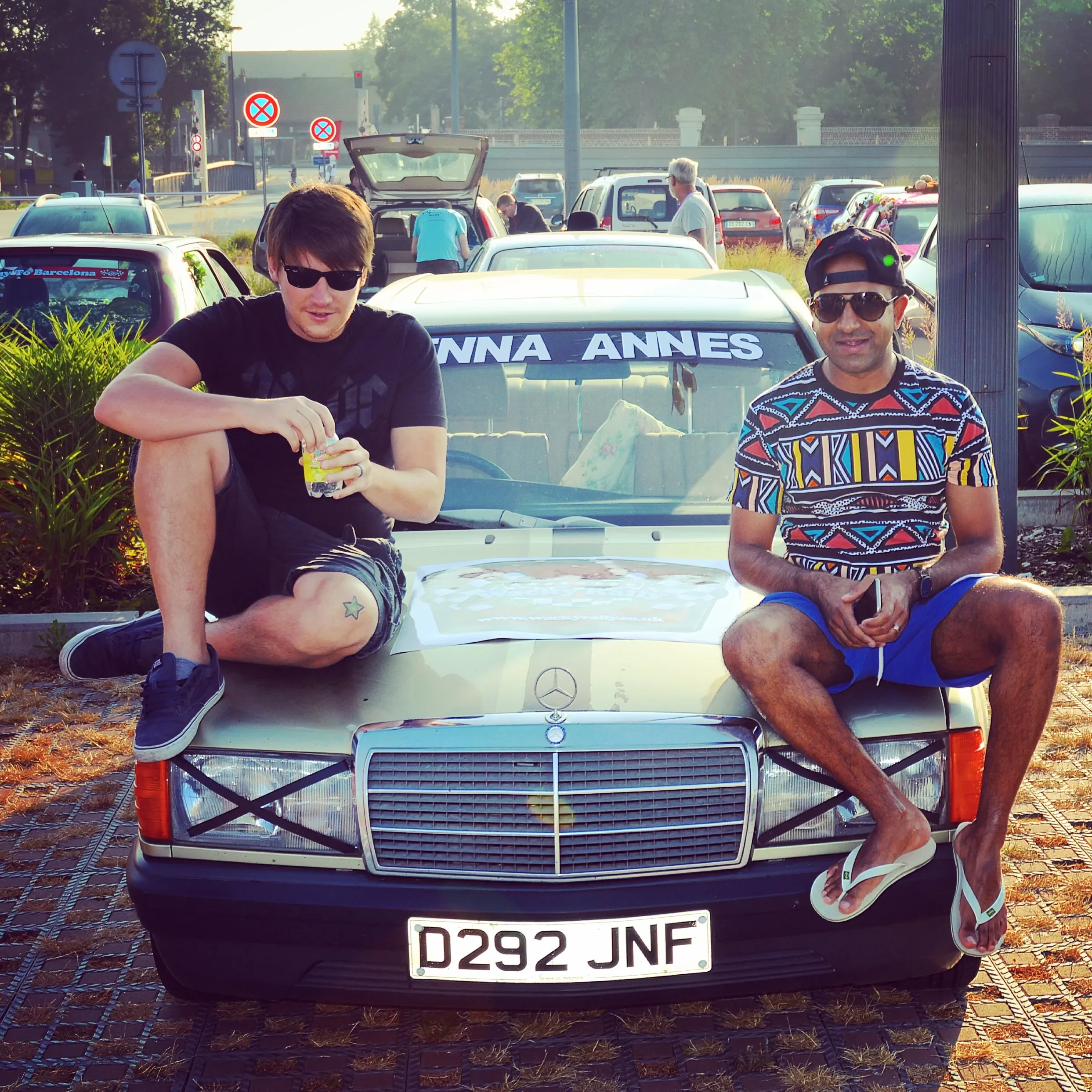 Two men sitting on the front hood of a vintage Mercedes-Benz car parked in a lot, with one wearing sunglasses and a black shirt, and the other wearing sunglasses, a colorful patterned shirt, and flip-flops. The background shows multiple parked cars a