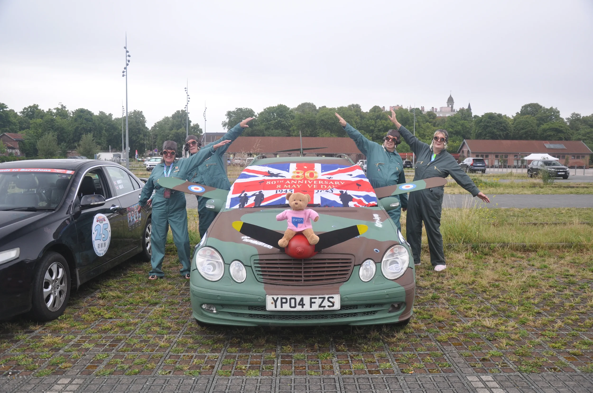 Group of four women in blue coveralls standing behind a car decorated with World War II aircraft images, a plush teddy bear, and a banner celebrating the 80th anniversary of VE Day, with other parked cars and a distant cityscape in the background.