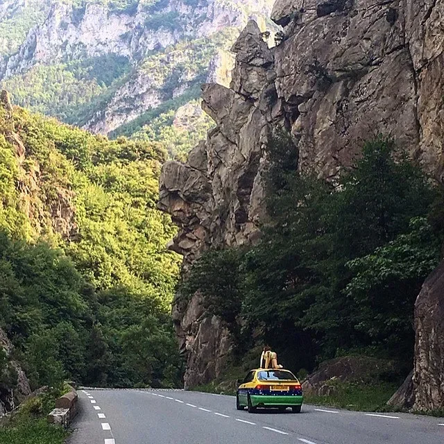 A car driving on a mountain road surrounded by lush green trees and large rocky cliffs.