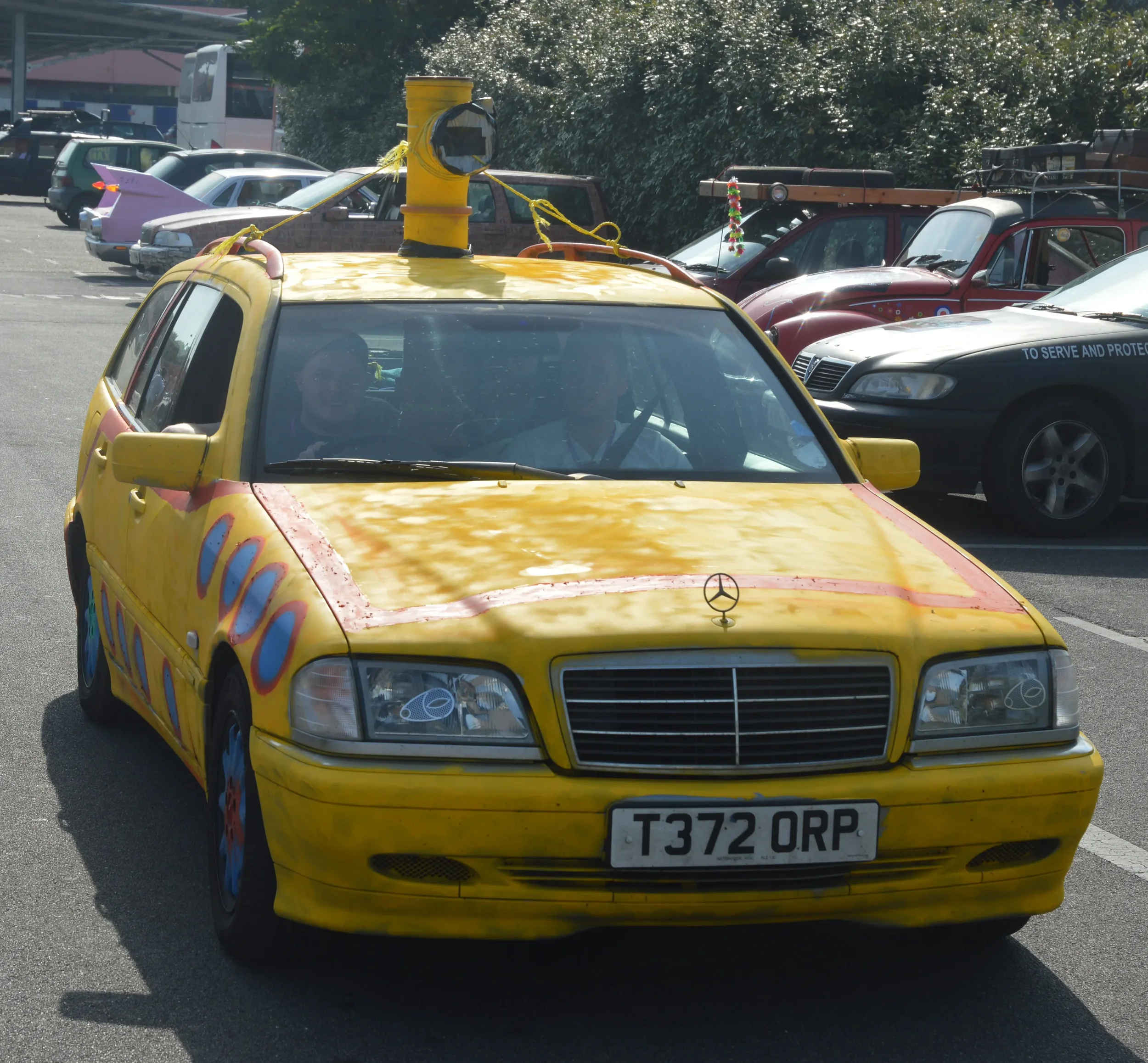 A yellow car with colorful swirls and a camera illustration on the front, resembling a mobile or media vehicle, parked in a lot with other cars around.