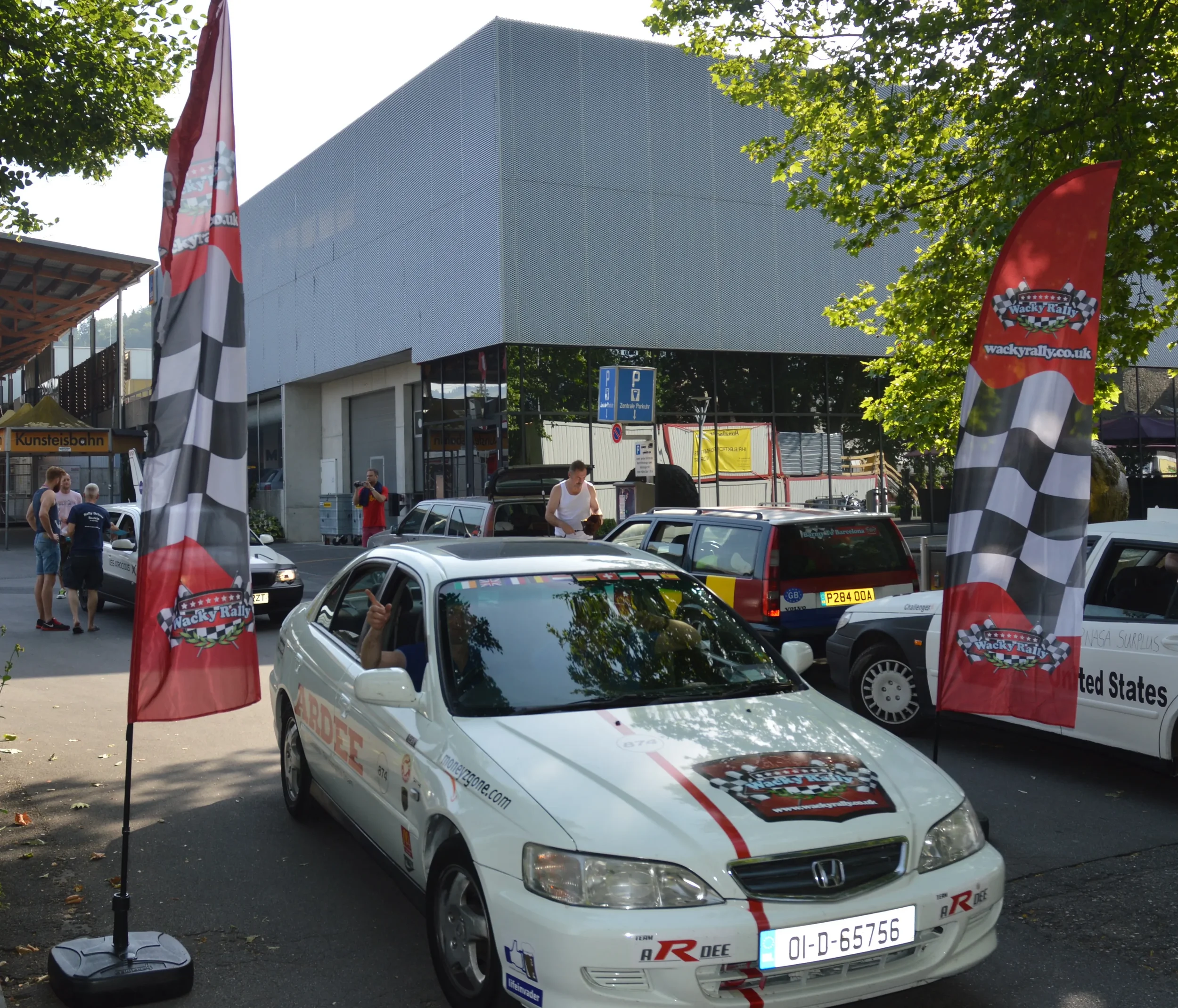 A white Honda car with rally stickers parked between two red and black checkered flags with 'Wacky Rally' logos; several other cars and people are visible in the background at a rally event, with trees and a modern building behind.