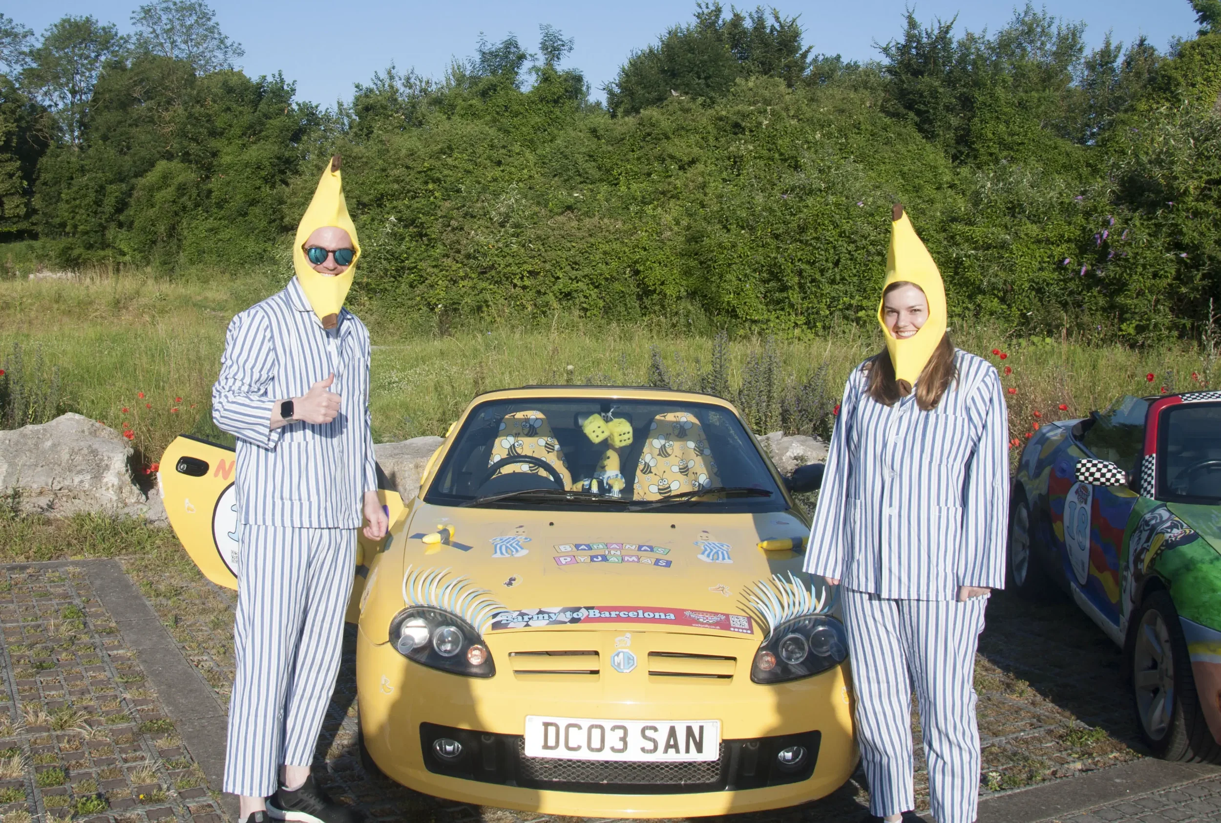 Two people dressed in matching pajamas and banana-shaped hats standing next to a yellow car decorated for a trip from Barns to Barcelona. They are outdoors with greenery in the background.