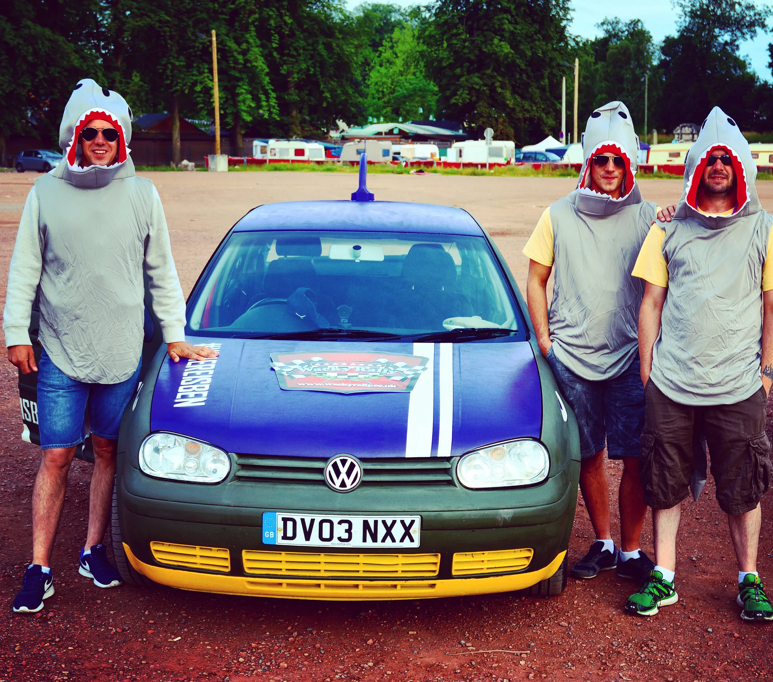 Four men wearing shark-themed hooded costumes standing around a Volkswagen car at an outdoor location with trees and caravans in the background.