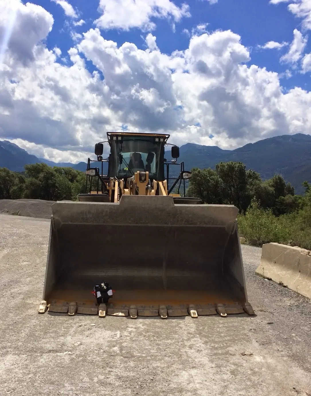 A large yellow construction tractor with a front loader bucket sits on dirt with mountains and partly cloudy sky in the background.