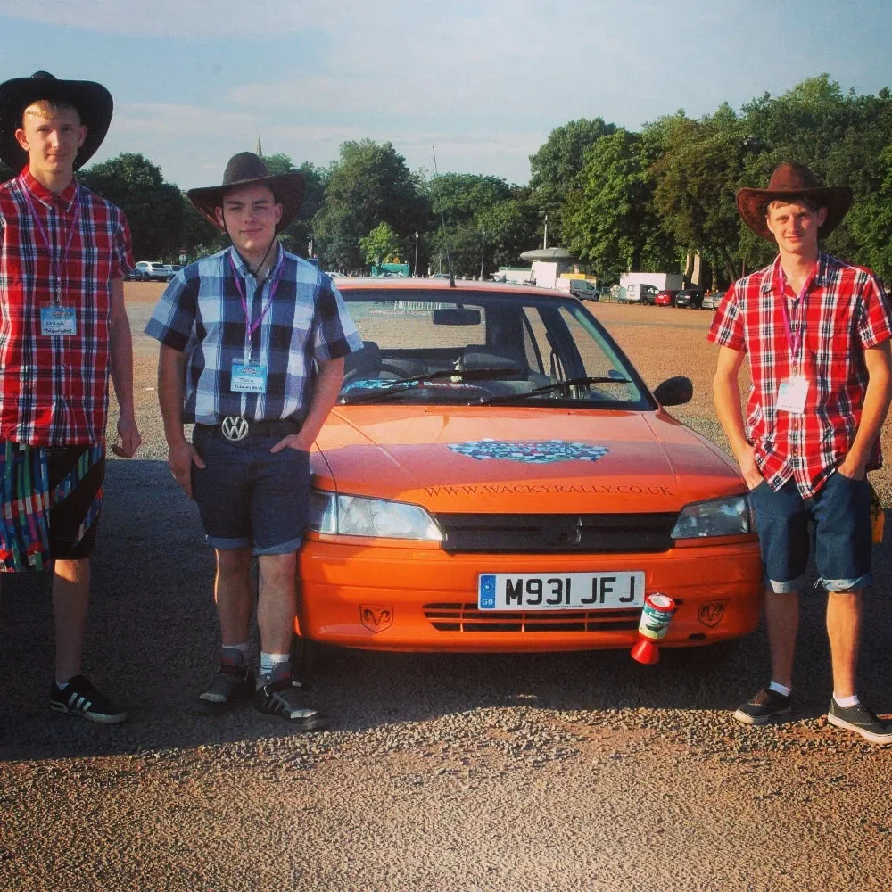 Four young men standing in front of an orange car with a website URL on the hood and a license plate M931 JFJ, all wearing plaid shirts and cowboy hats, in an outdoor parking lot.