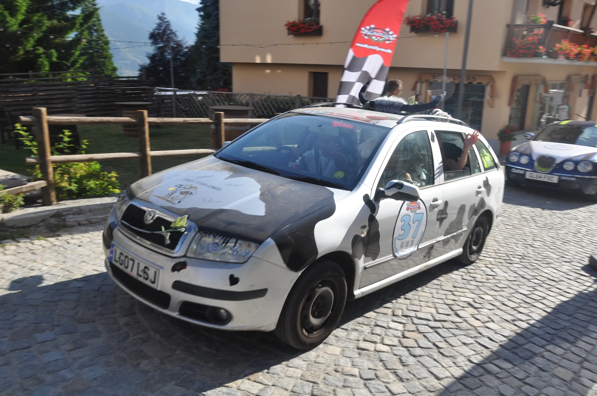 A decorated car participating in a rally, featuring racing stickers, a number 37, a cow print design, and a roof-mounted bull horn. The car is on a cobblestone street with buildings and people in the background.