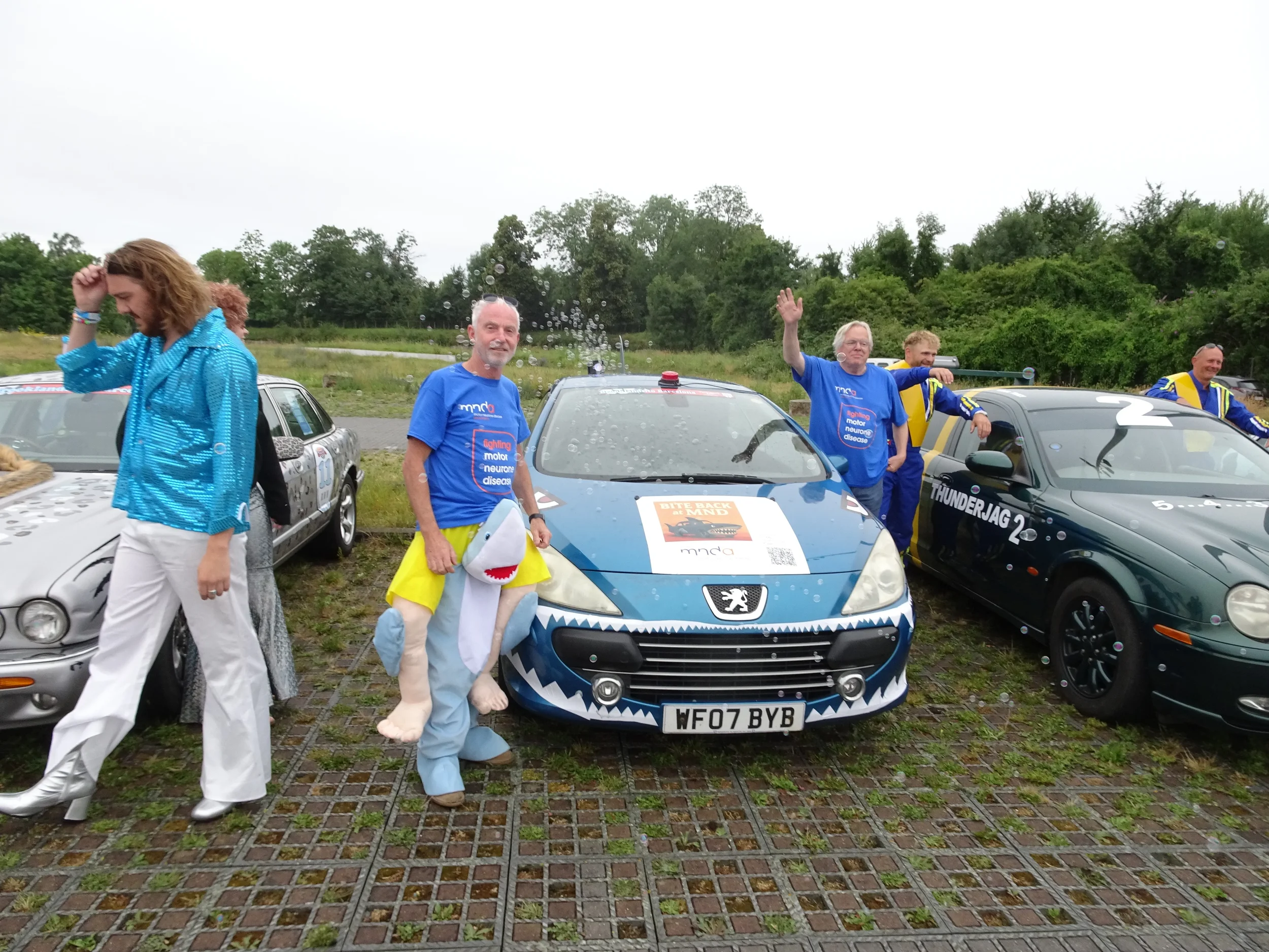 Group of people standing between customized cars during outdoor event, some wearing blue shirts, one person dressed in shark costume, overcast sky, grassy field in background.
