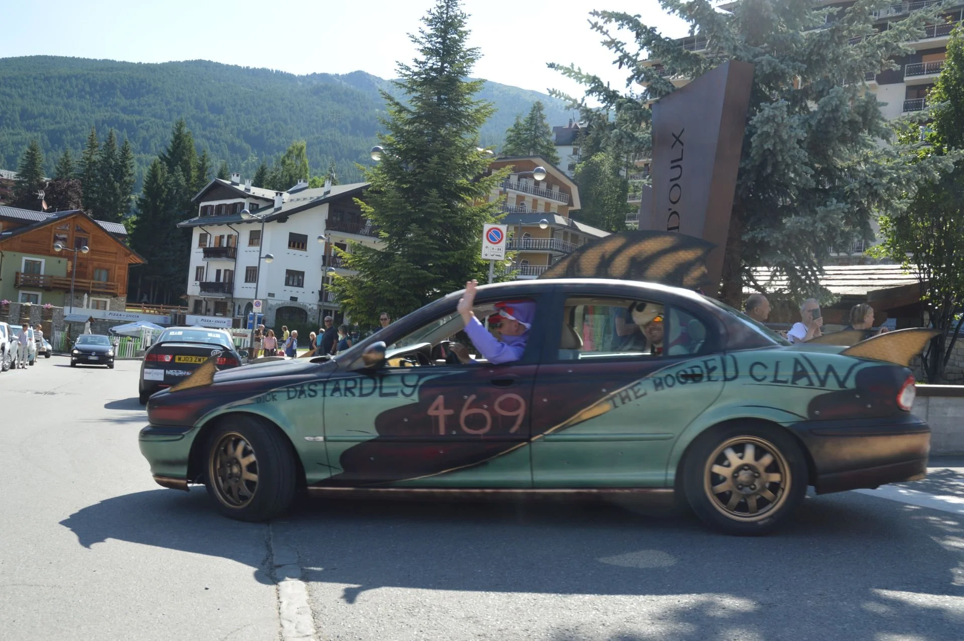 A colorful car decorated with artwork and the names 'Dick Dastardly' and 'The Hooded Claw' is driving through a mountain town with buildings, trees, and people in the background.