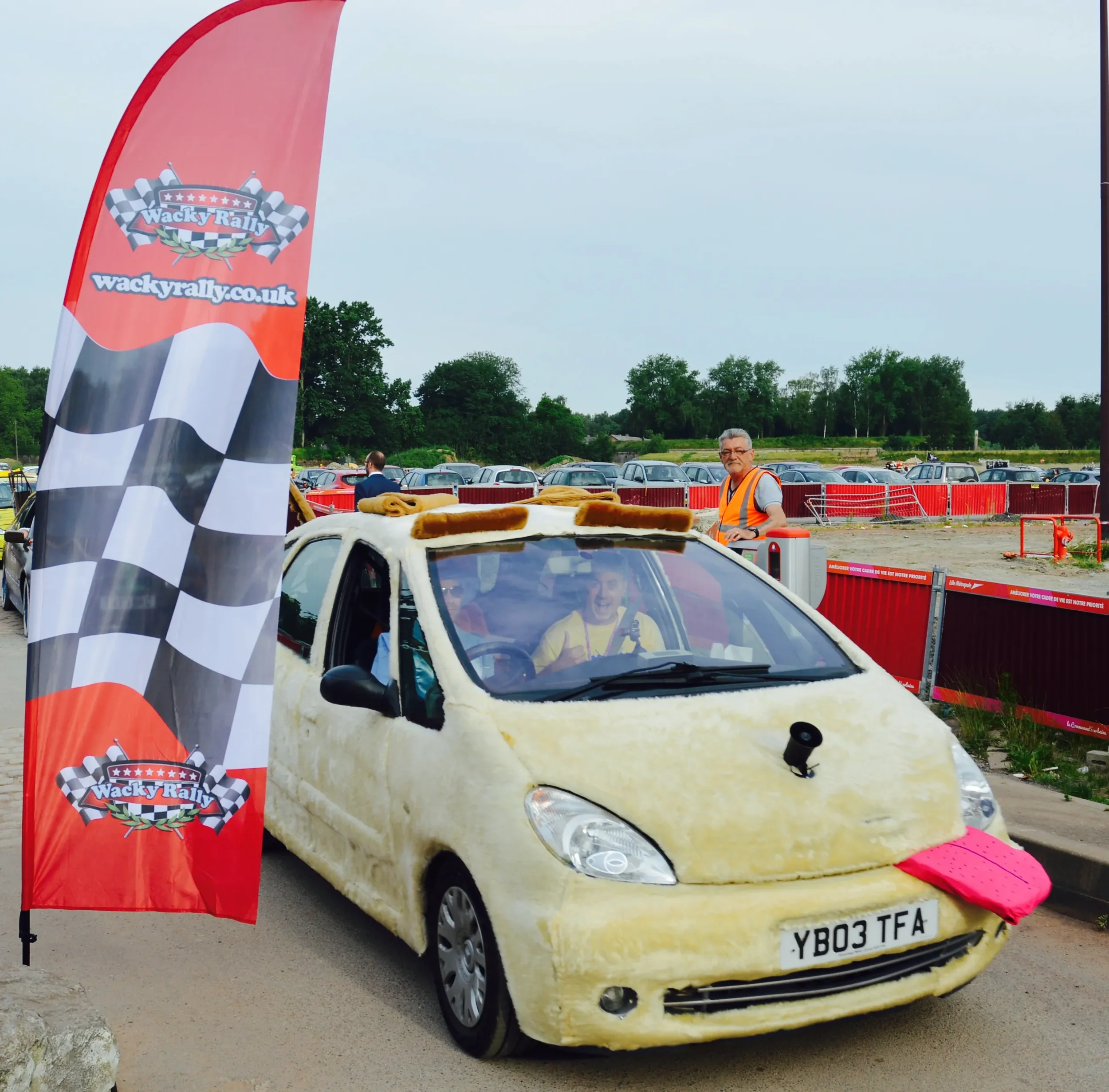 A small car covered in fluffy, yellow material to resemble a fried egg, with a pink object as a yolk on the front. The car is parked next to a red and black checkered flag banner with rally logos. Two people are inside and one person is standing outs