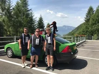 Four men standing next to a green and black car on a mountain road with trees and mountains in the background.