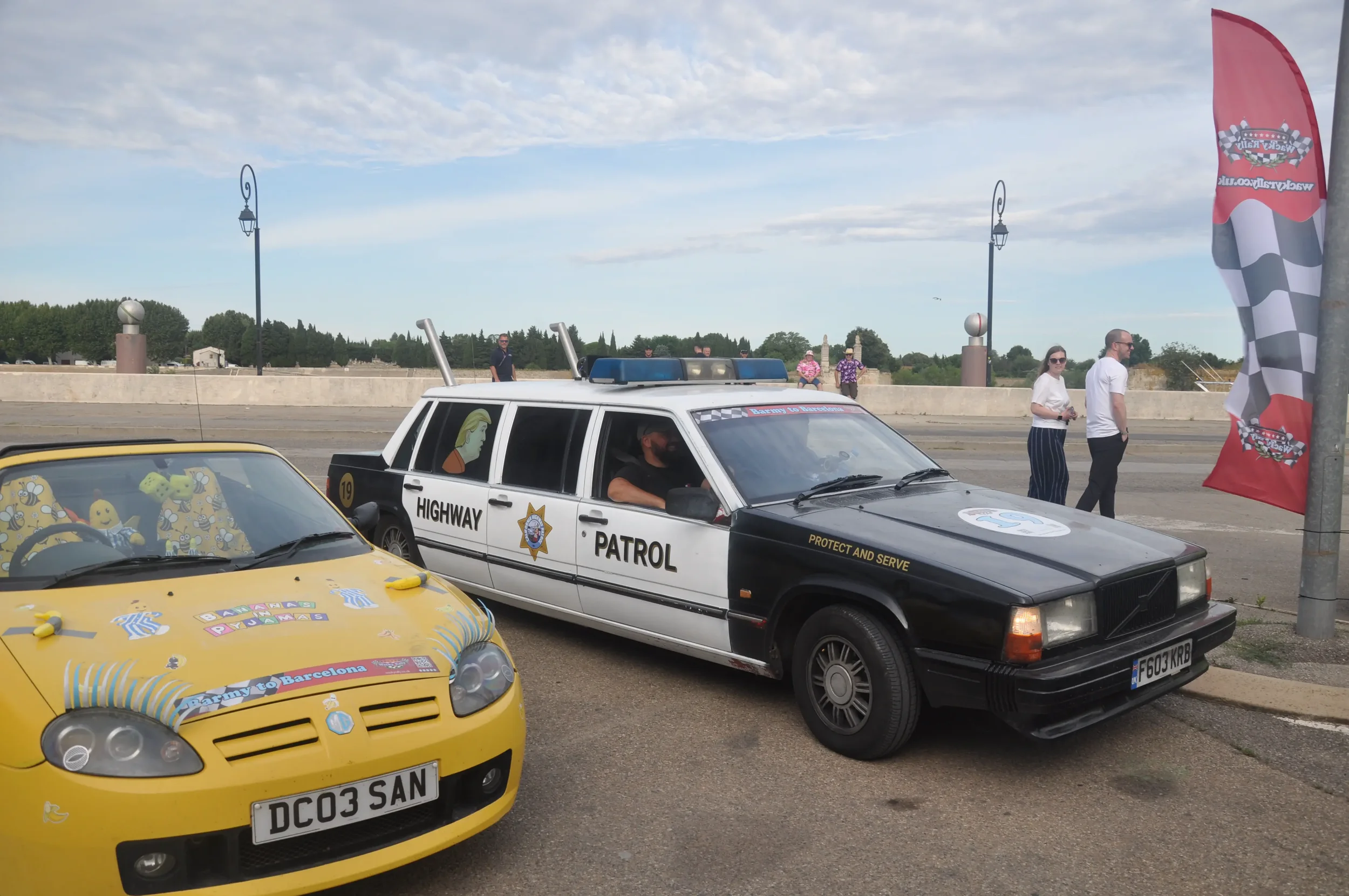A police car labeled 'Highway Patrol' and a yellow convertible car with decorations and writings on the hood, parked outdoors during daytime with a few people standing nearby.