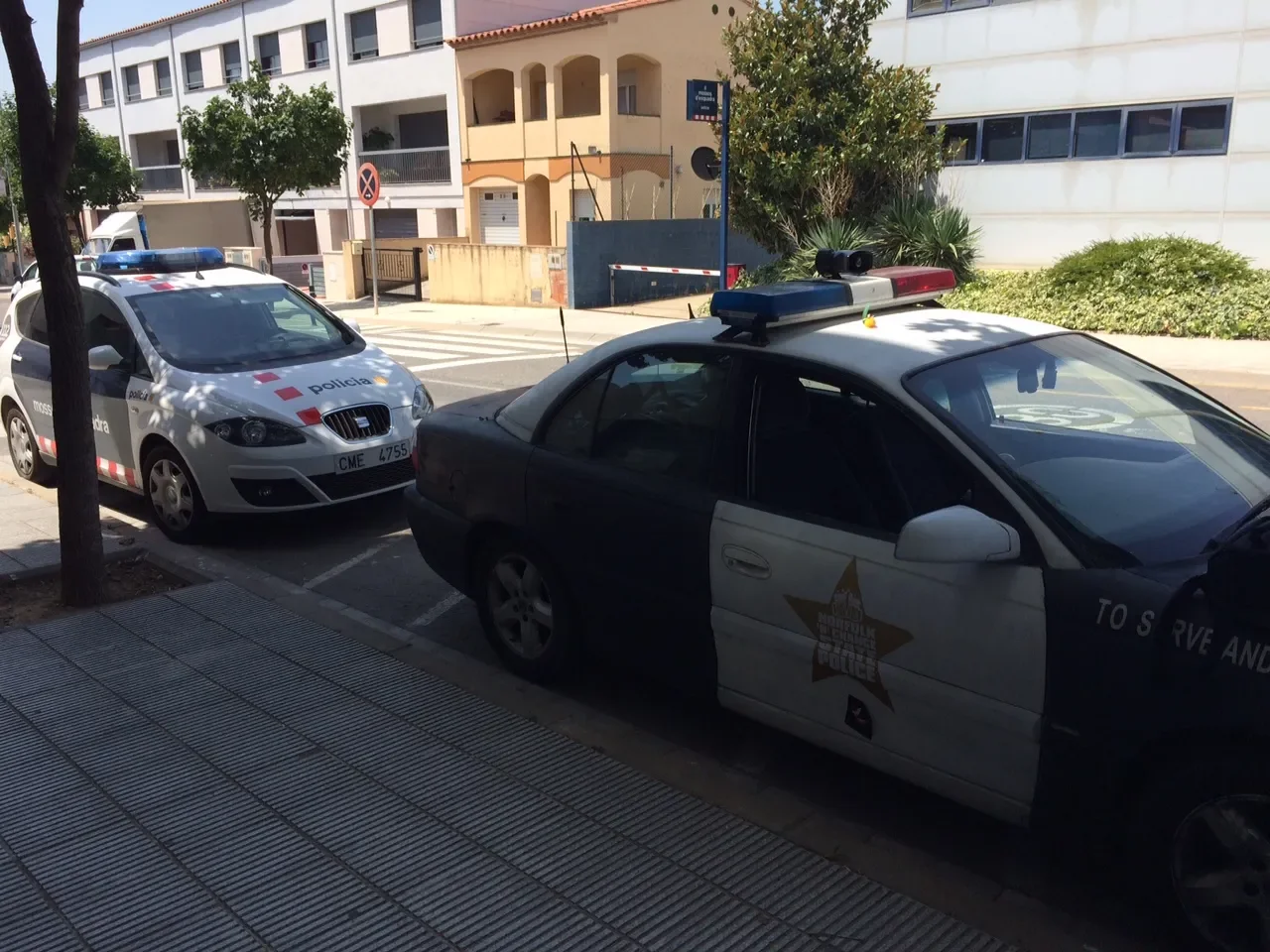 Two police cars parked on the side of a city street with residential buildings and trees in the background.