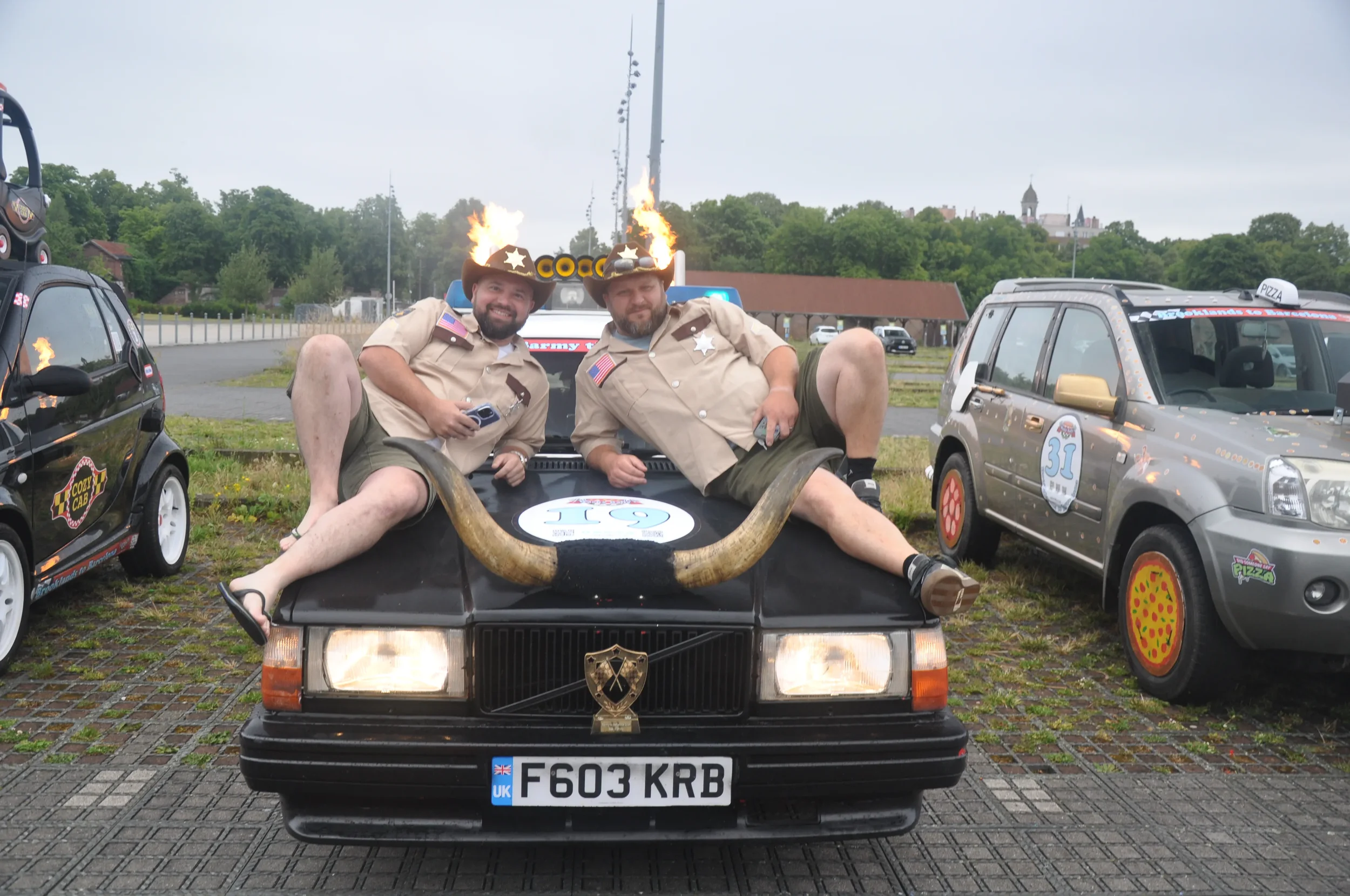 Two men in sheriff costumes sitting on the hood of a black car with a large Goodyear tire bullhorn, surrounded by decorated toy cars, some with fire effects and themed accessories, in an outdoor parking lot.