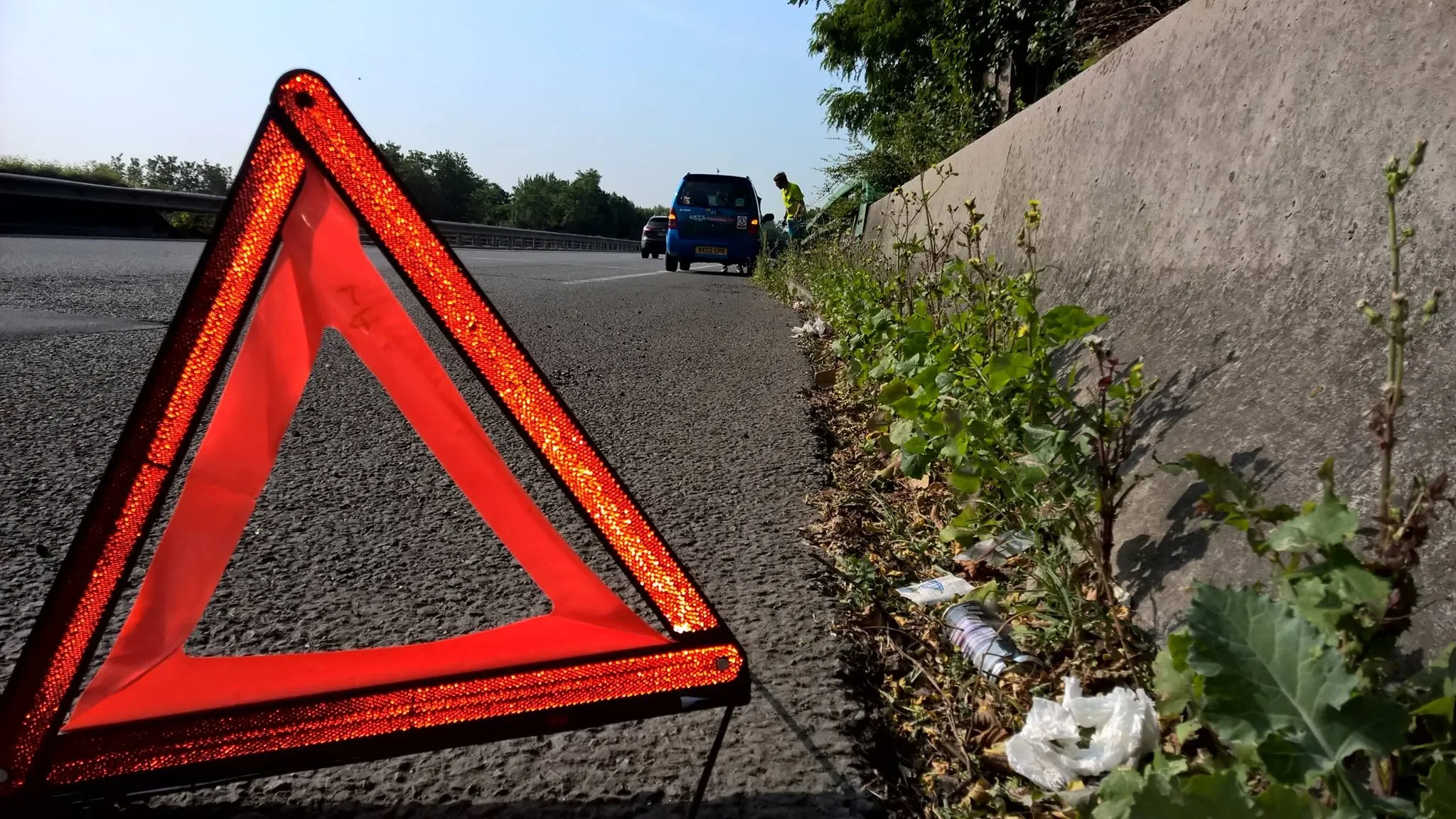 A red reflective warning triangle on the side of a road near some trash and plants. Several vehicles and a person are in the background.
