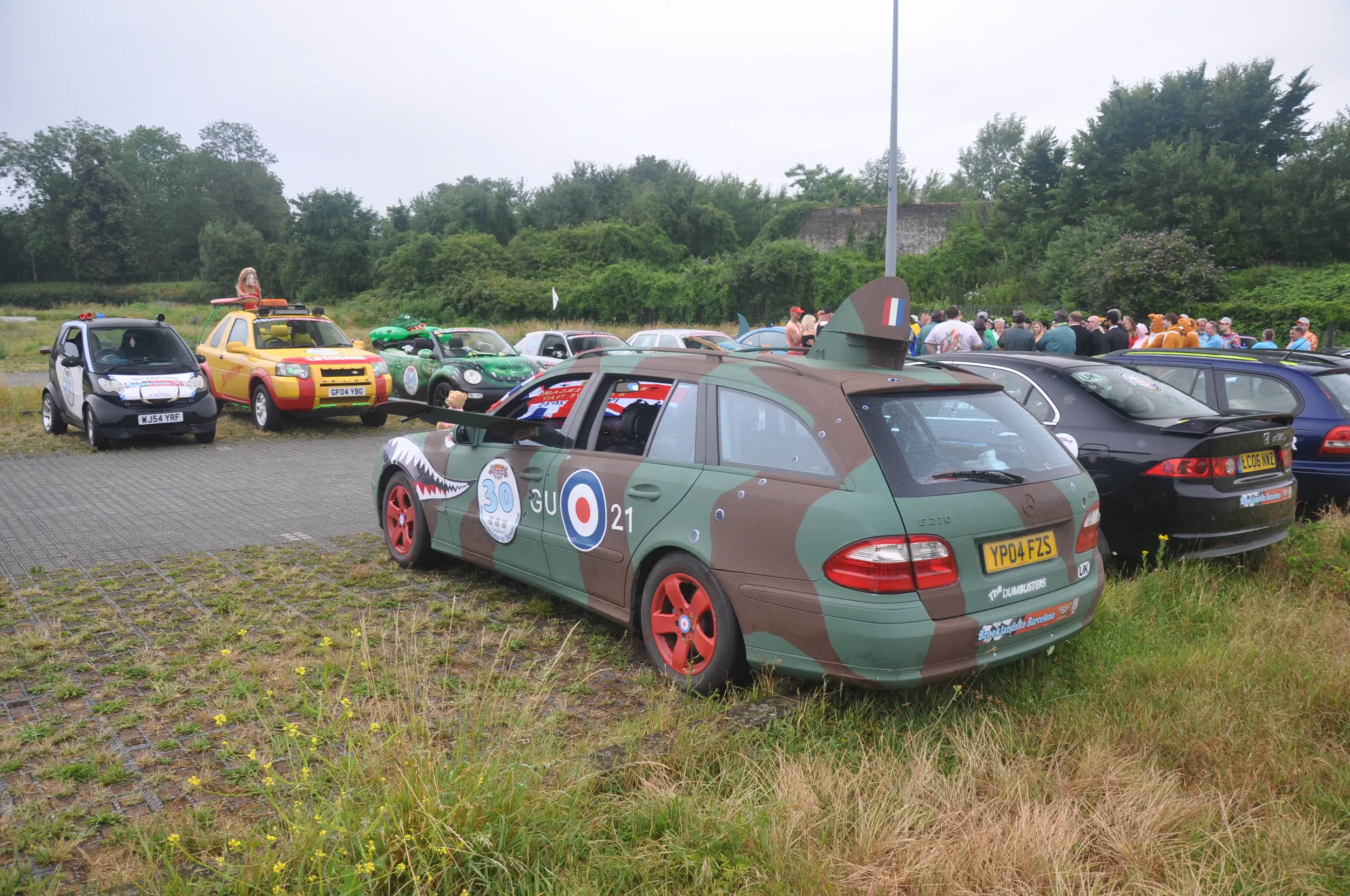Car decorated with military camouflage design and cartoon USS Enterprise nose art, parked in a lot with other vehicles, and a group of people in the background