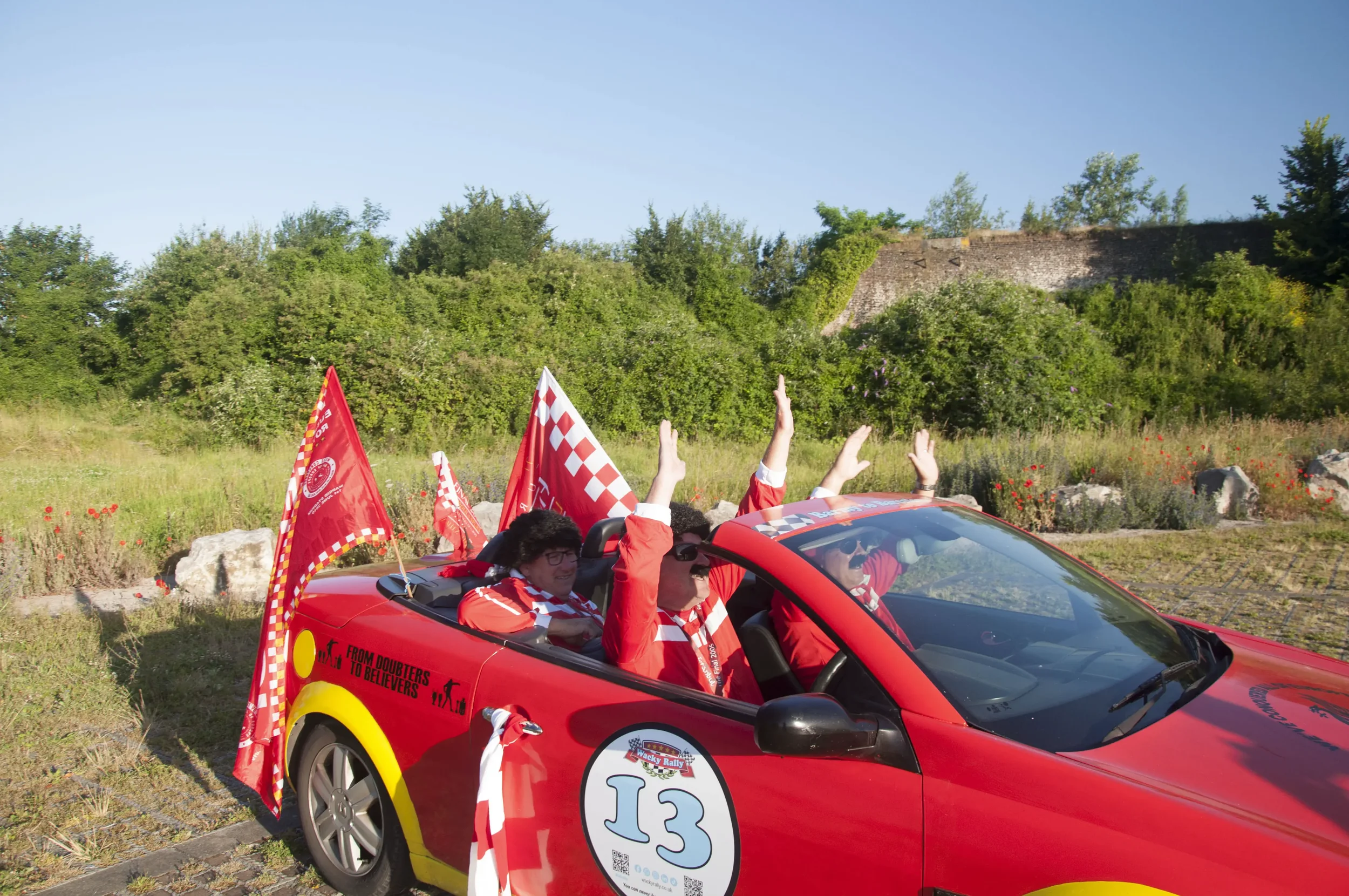 Group of people in a red convertible car celebrating, waving hands, with checkered flags and rally stickers, in a grassy area with trees and rocks in the background.