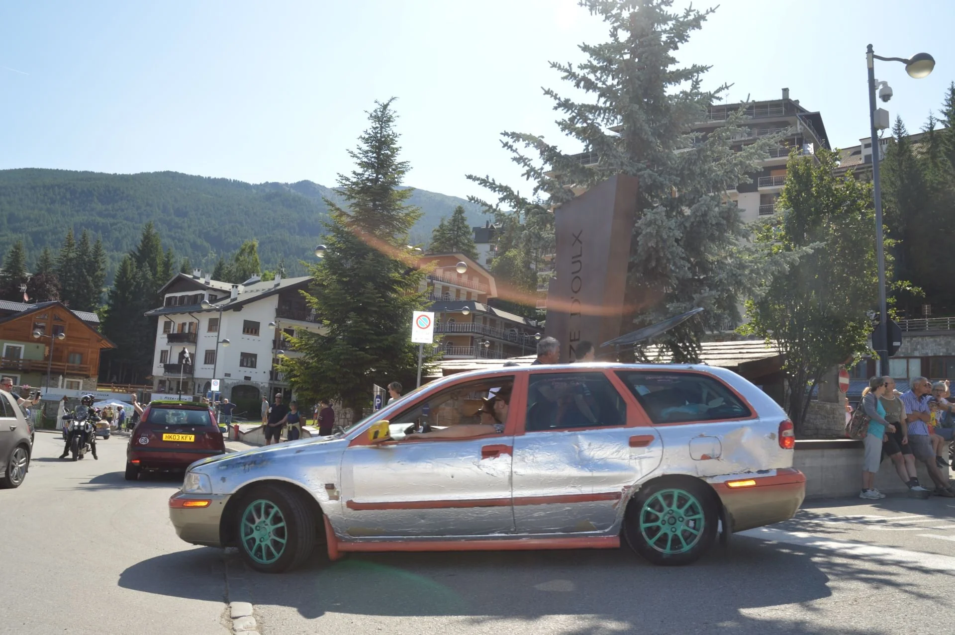 A silver station wagon with orange accents and decals, parked in a busy outdoor area with people walking and standing around. The background features trees, buildings, and a mountain landscape underneath a clear, sunny sky.