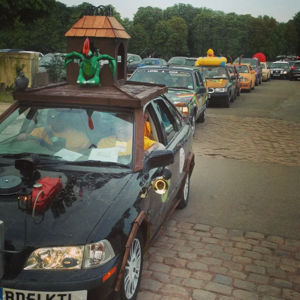 Decorated cars lined up in a parade, with the front car featuring a dragon sculpture on the roof and props on the side, in a parking area with trees in the background.