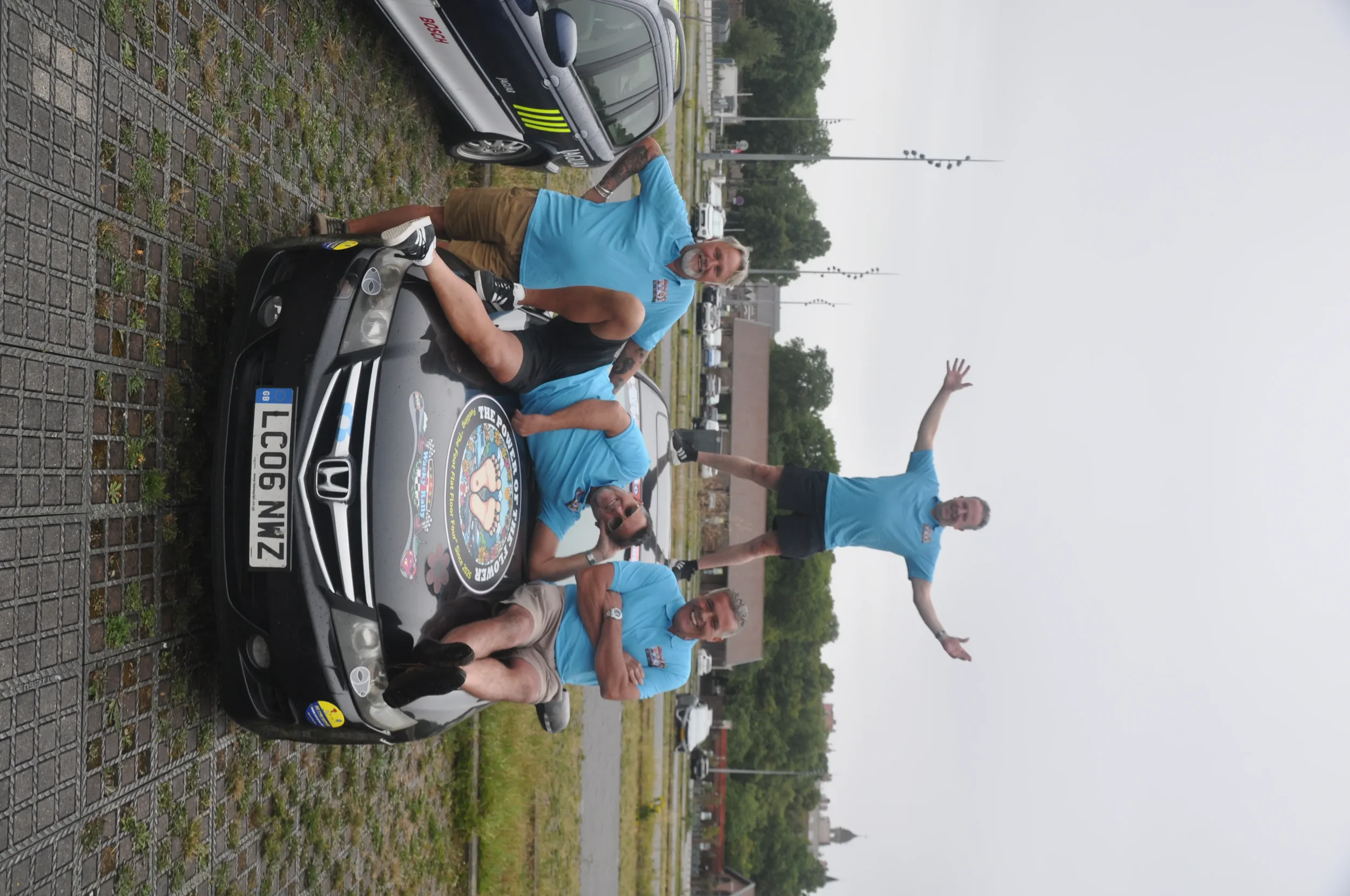 Four men wearing matching light blue shirts are gathered around a black sports car with a sticker on the hood, parked on a cobblestone surface. One man is lying on the car, another is sitting with his legs crossed, another is standing with his arms c