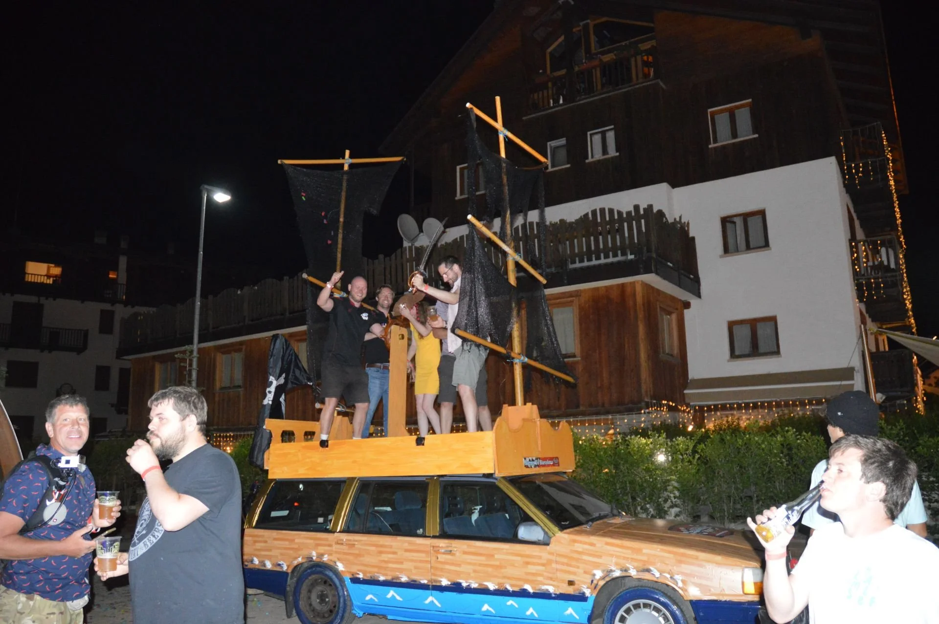 People celebrating on a decorated car with a boat mast structure, at night in front of a modern building, with some holding drinks and wearing party attire.