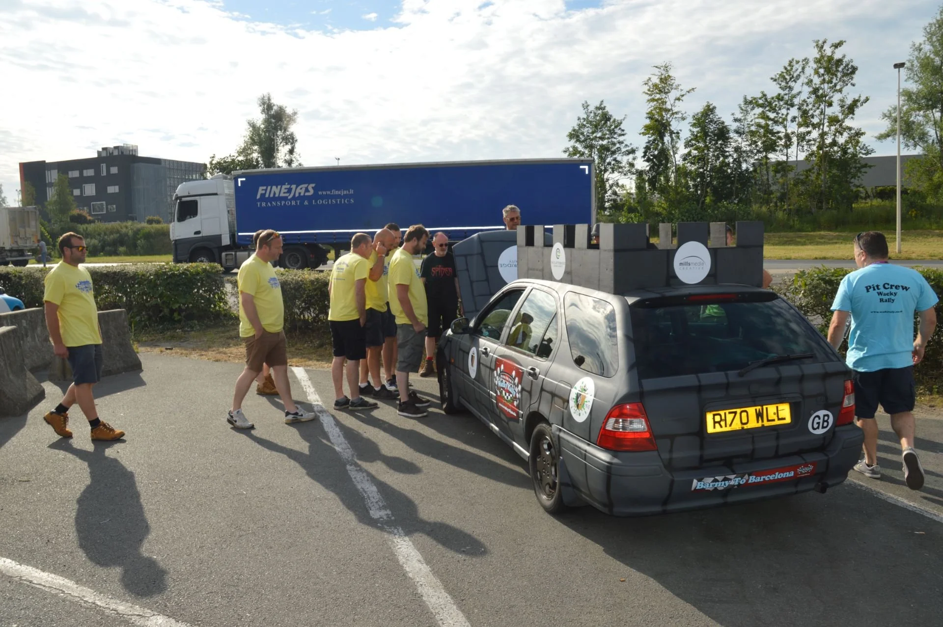 Group of men gathered around a small, decorated car parked in a lot, with a large blue truck in the background. The car has various stickers and a roof load.