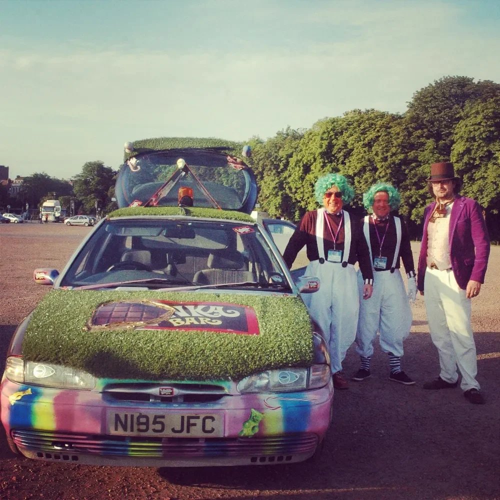Three people dressed in colorful, whimsical costumes standing beside a car decorated with fake grass, rainbow colors, and playful designs, parked in an open area with trees in the background.