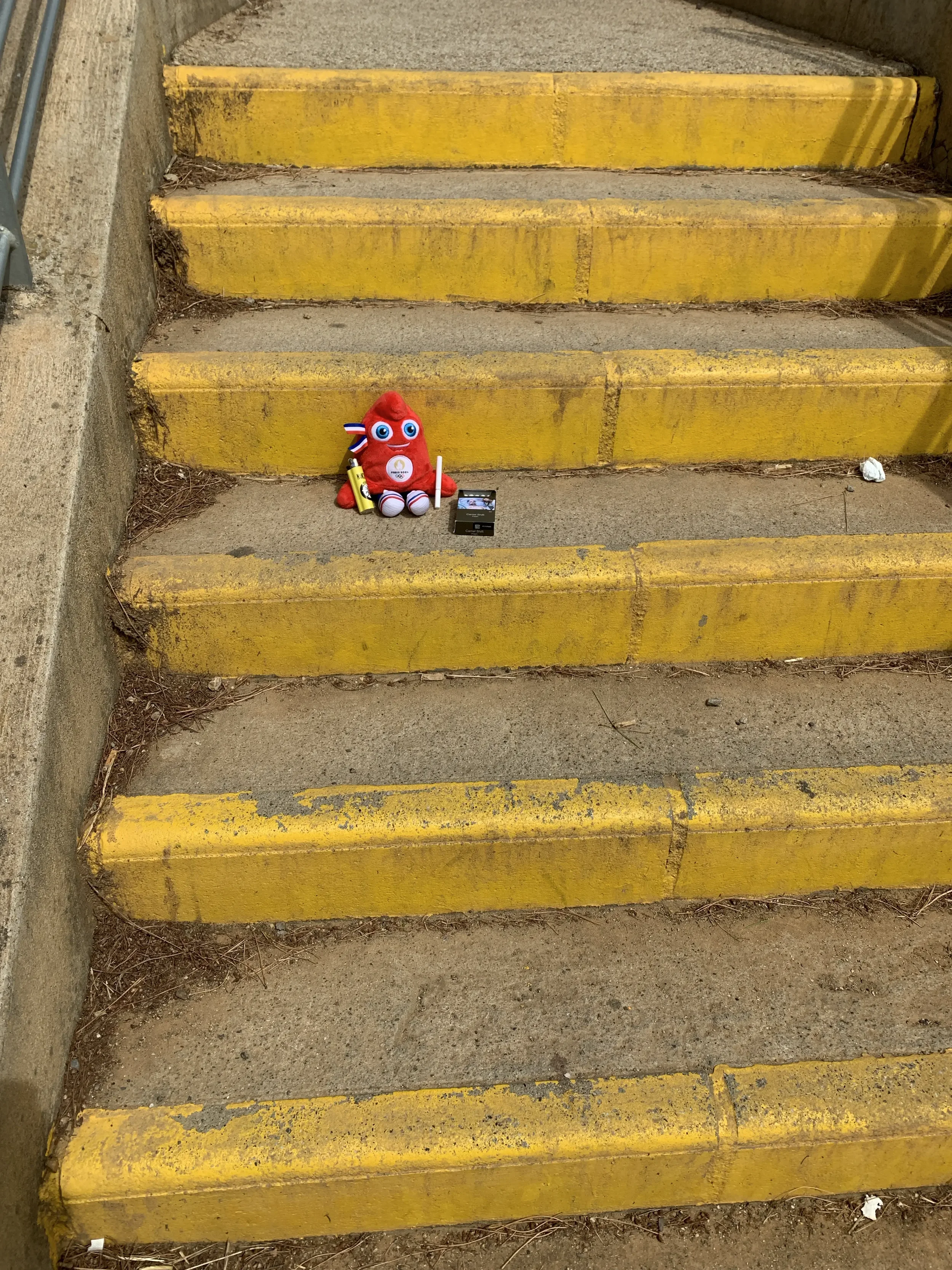 A small red plush toy with googly eyes, holding a yellow object, sits on concrete stairs painted yellow. There is a small black box and a white object nearby, with dirt and debris surrounding the stairs.