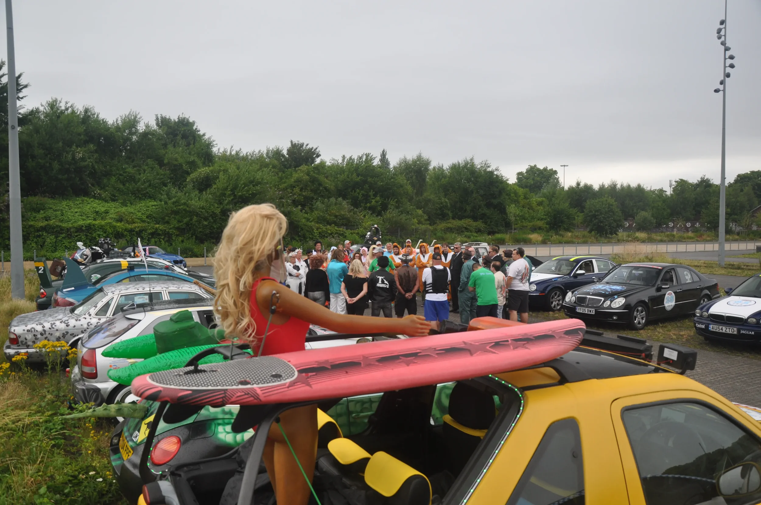 Person with long blonde hair standing on a yellow car, holding a pink surfboard, in a parking lot filled with cars and a crowd of people gathered around.