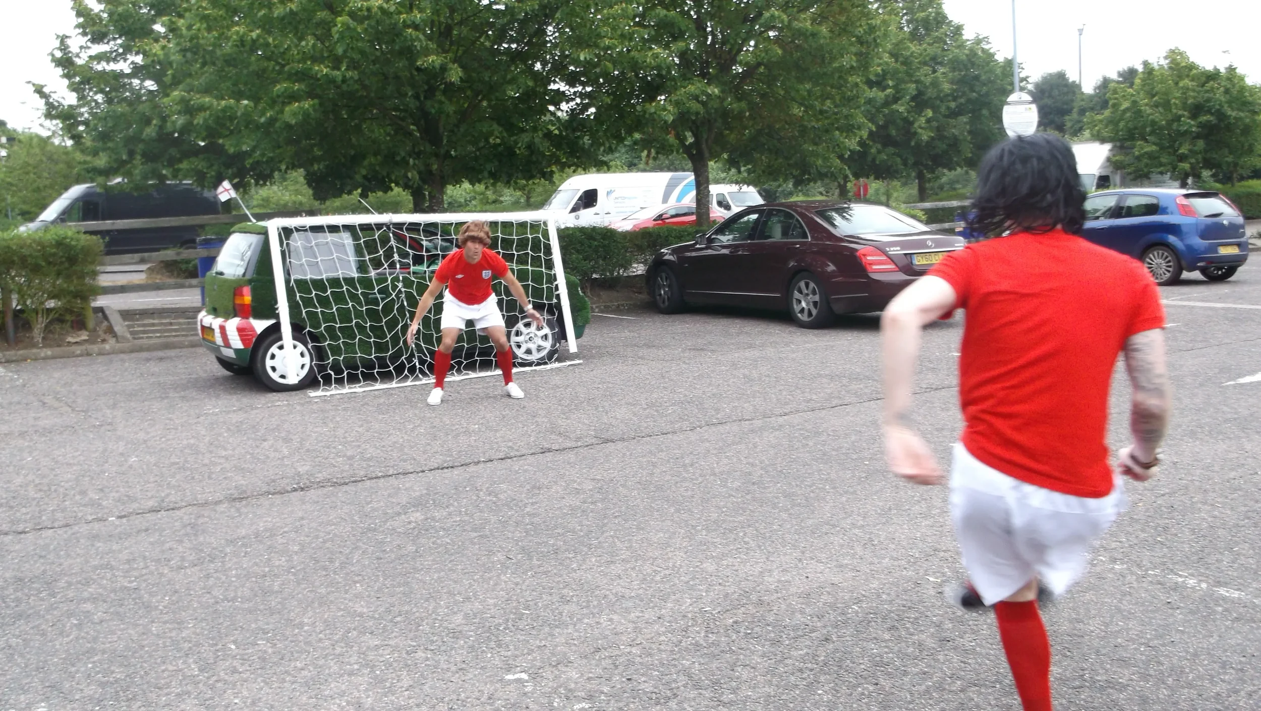 Two soccer players in red jerseys practicing shooting on goal in a parking lot, with cars and trees in the background.