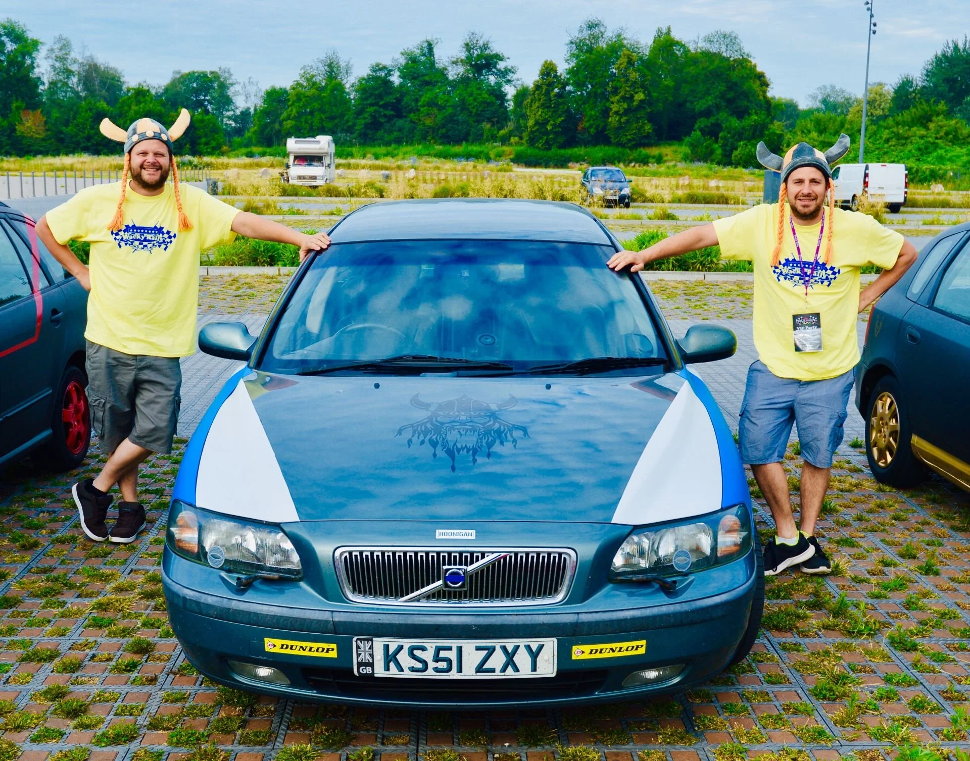 Two men wearing Vikings helmets and yellow shirts leaning on a black and silver Volvo car in a parking lot with grassy areas and trees in the background.
