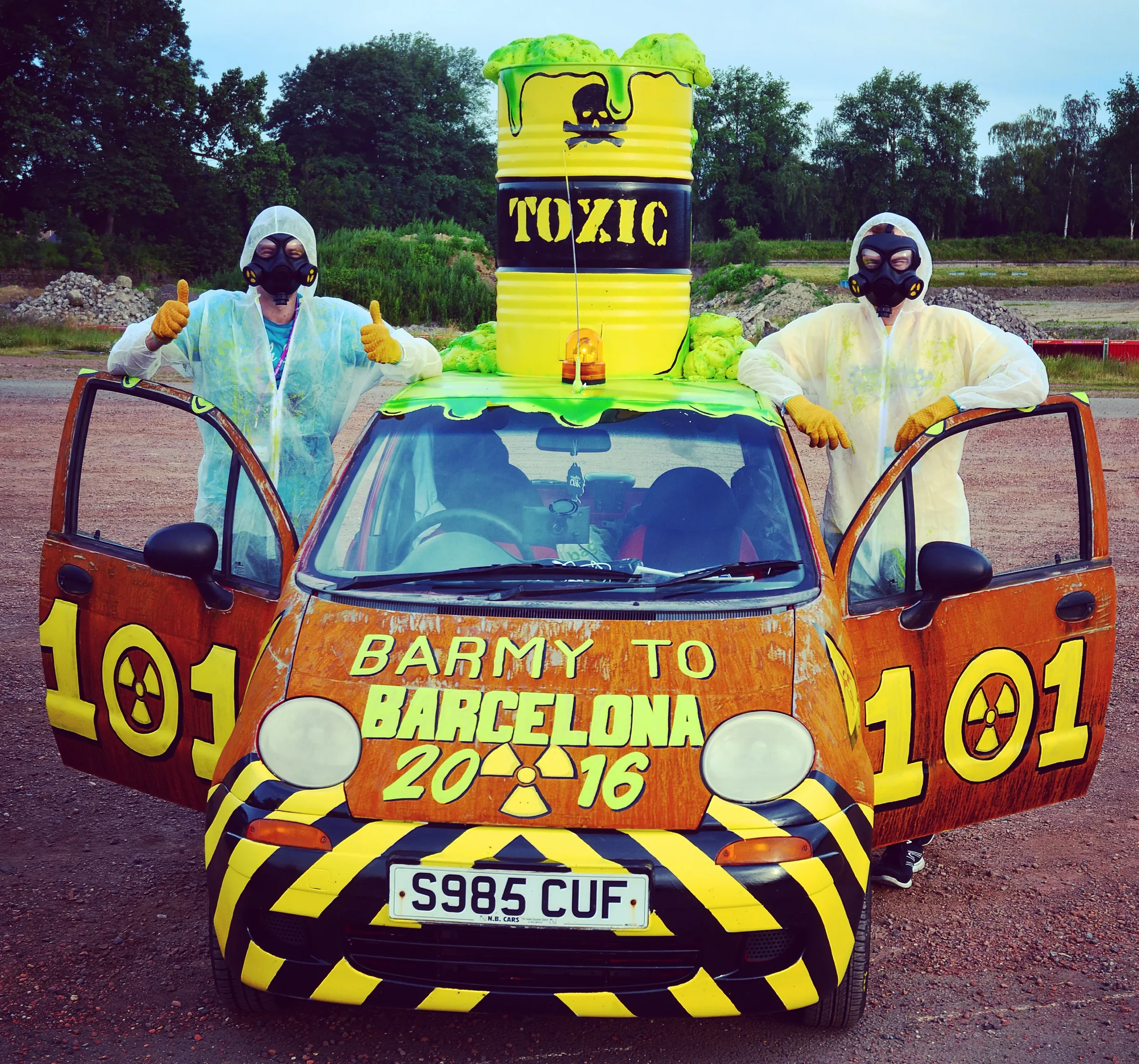 Two people wearing hazmat suits and gas masks standing beside a car painted with radioactive symbols and the text 'Barmy to Barcelona 2016'. The car has a large container on its roof labeled 'Toxic' with a skull and crossbones symbol, resembling a ba