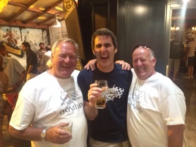 Three men smiling and posing for a photo in a lively bar or pub. The man in the middle is holding a glass, and all are wearing casual shirts.