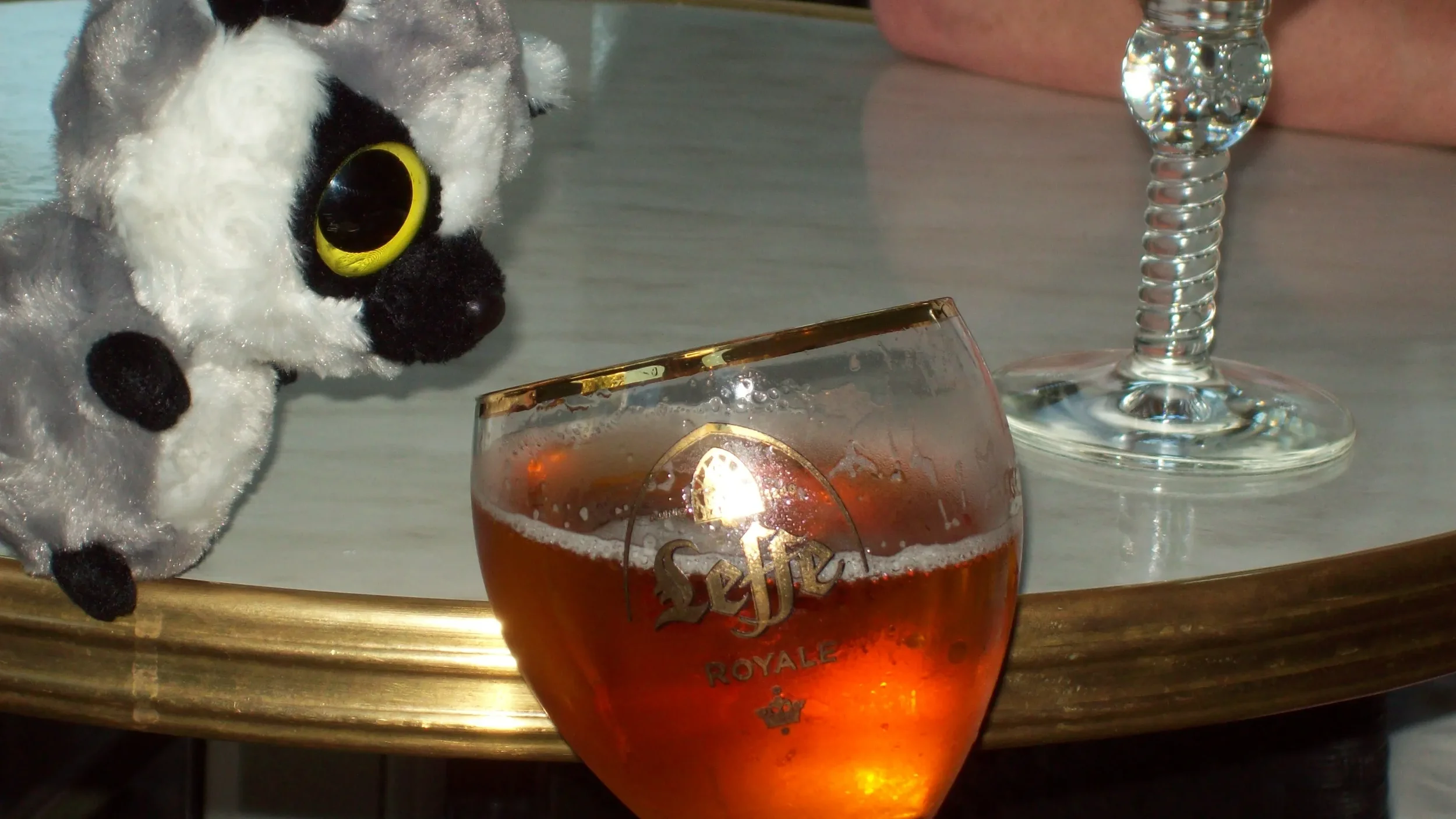 A plush toy dog with large yellow eyes looking at a glass of beer on a table, with a wine glass and part of a person's arm in the background.