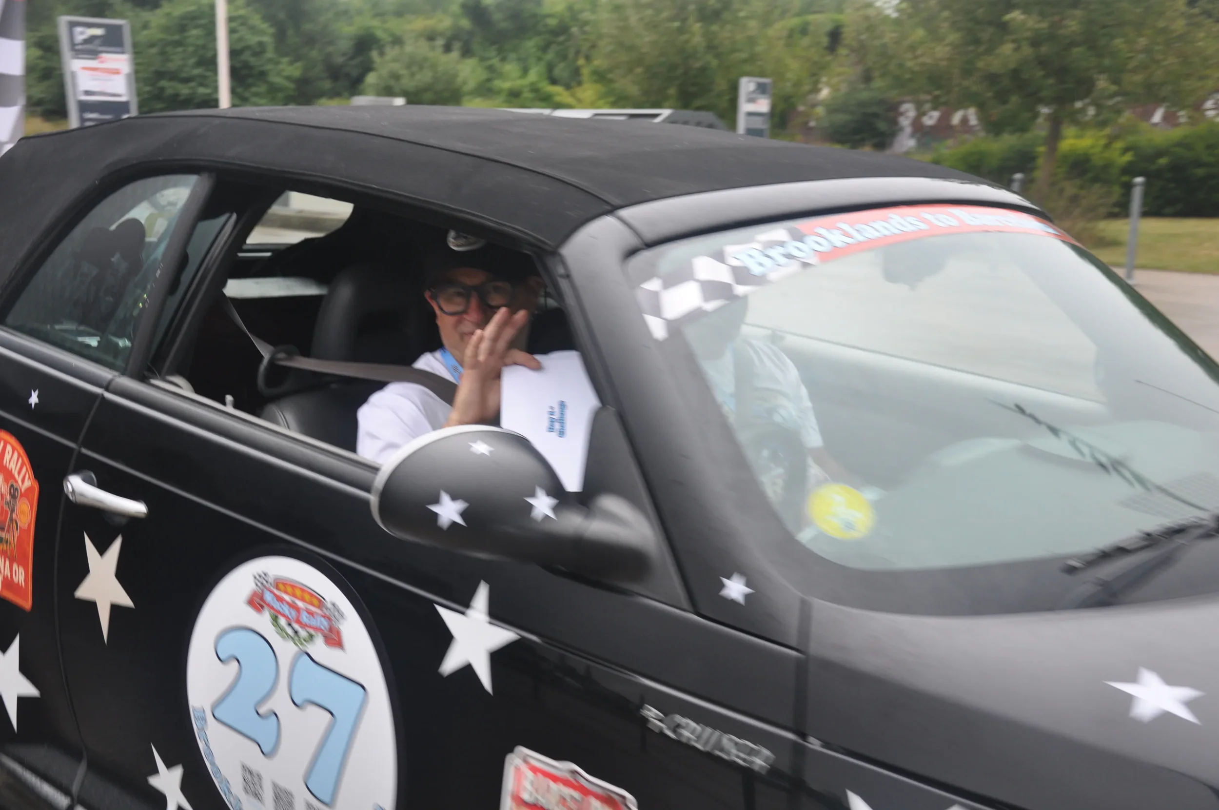 A man in a racing car waving and holding a piece of paper, wearing glasses, black cap, and a white shirt. The car is decorated with stars and race stickers, with the number 27 on the side.