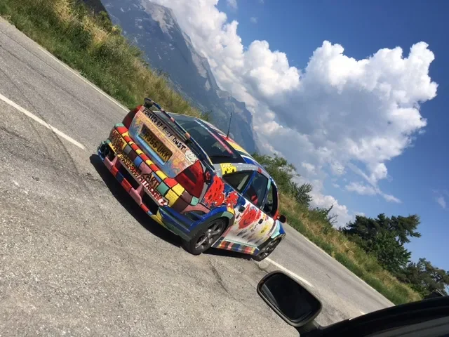 Colorful car with artwork and stickers on the side, parked on the side of a rural road with mountains and blue sky in the background.