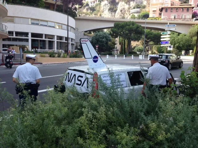 Two police officers in uniform standing near a small model spacecraft with NASA and SpaceX logos, placed on a patch of greenery beside a street in an urban area.