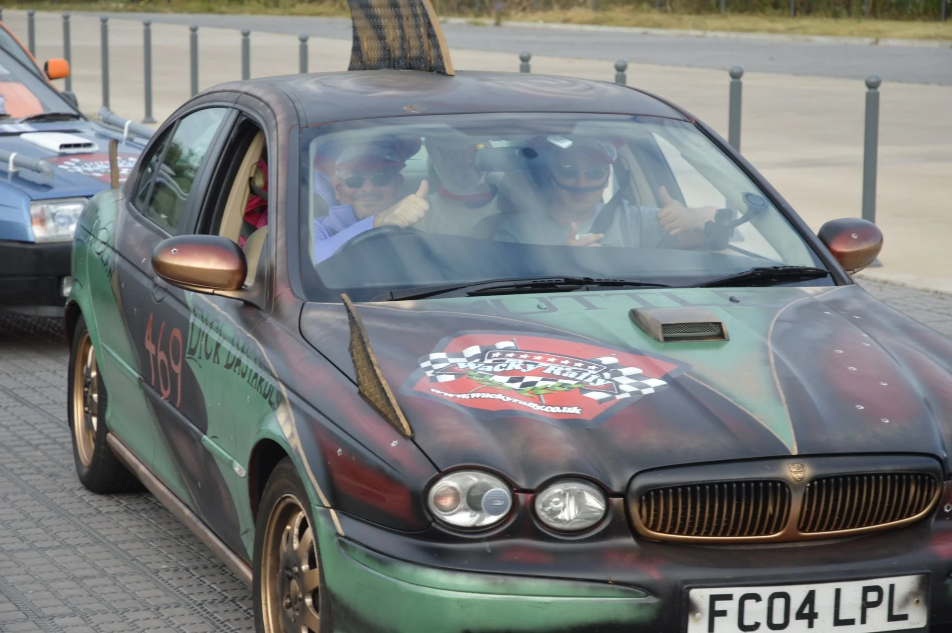 A group of four people in a decorated rally car, giving thumbs up and smiling, parked on a paved surface with a fence in the background.