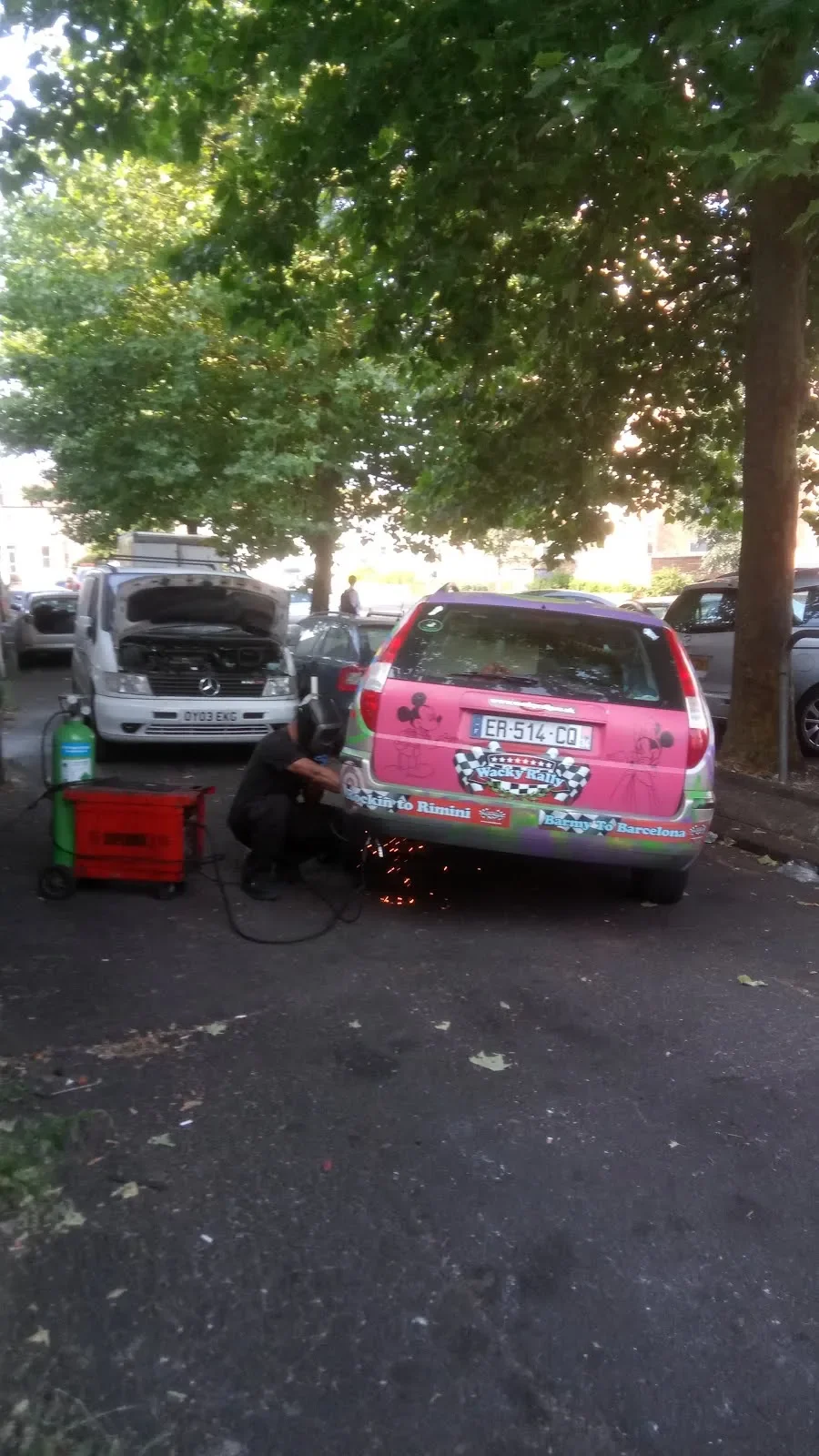 A person welding the back of a pink car decorated with racing-themed stickers, outdoors near trees and parked cars, with a welding mask and sparks visible.