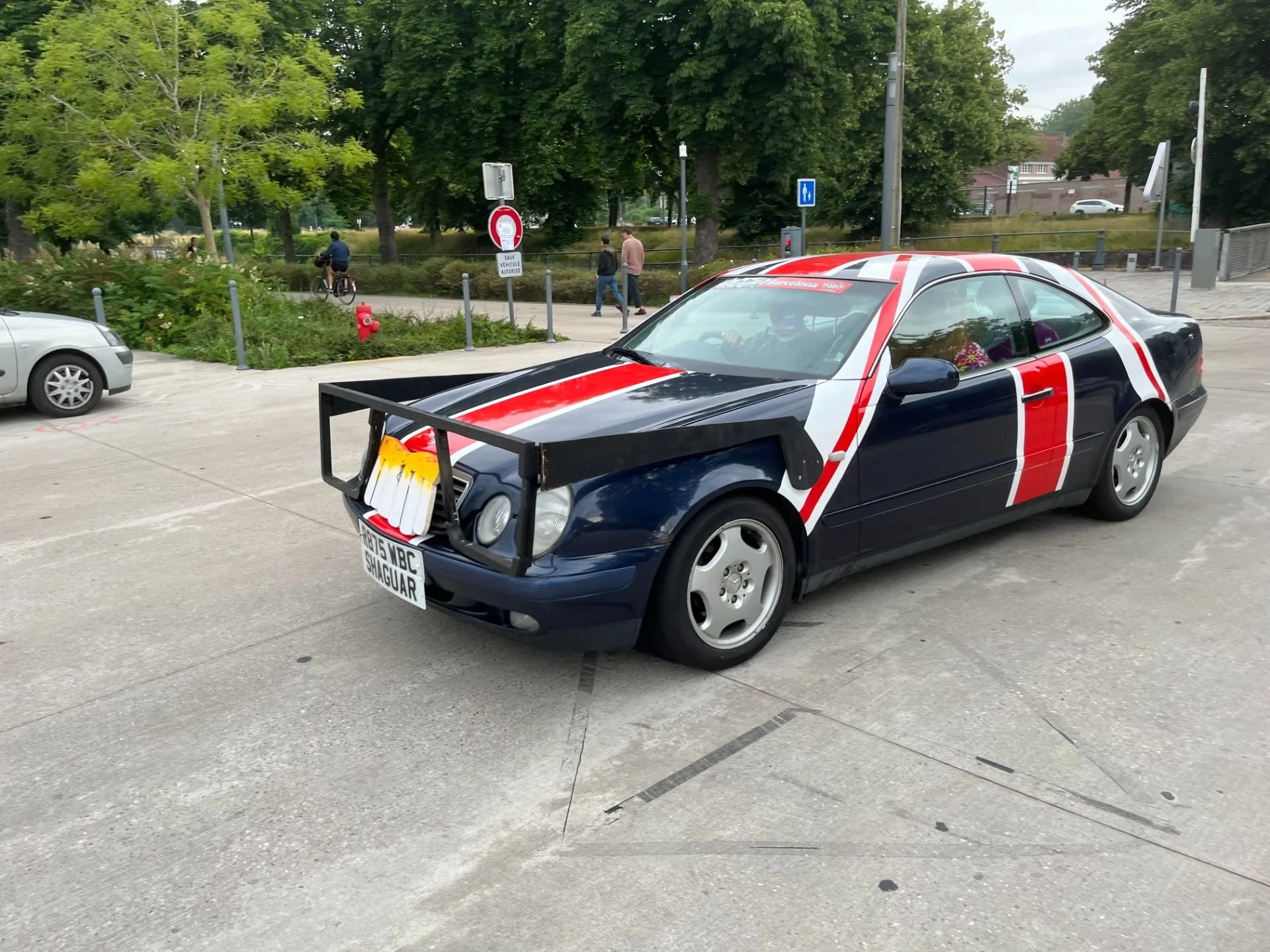 A modified black and red sports car with a front bumper guard and custom paint design parked in a parking lot. There are trees and pedestrians in the background.