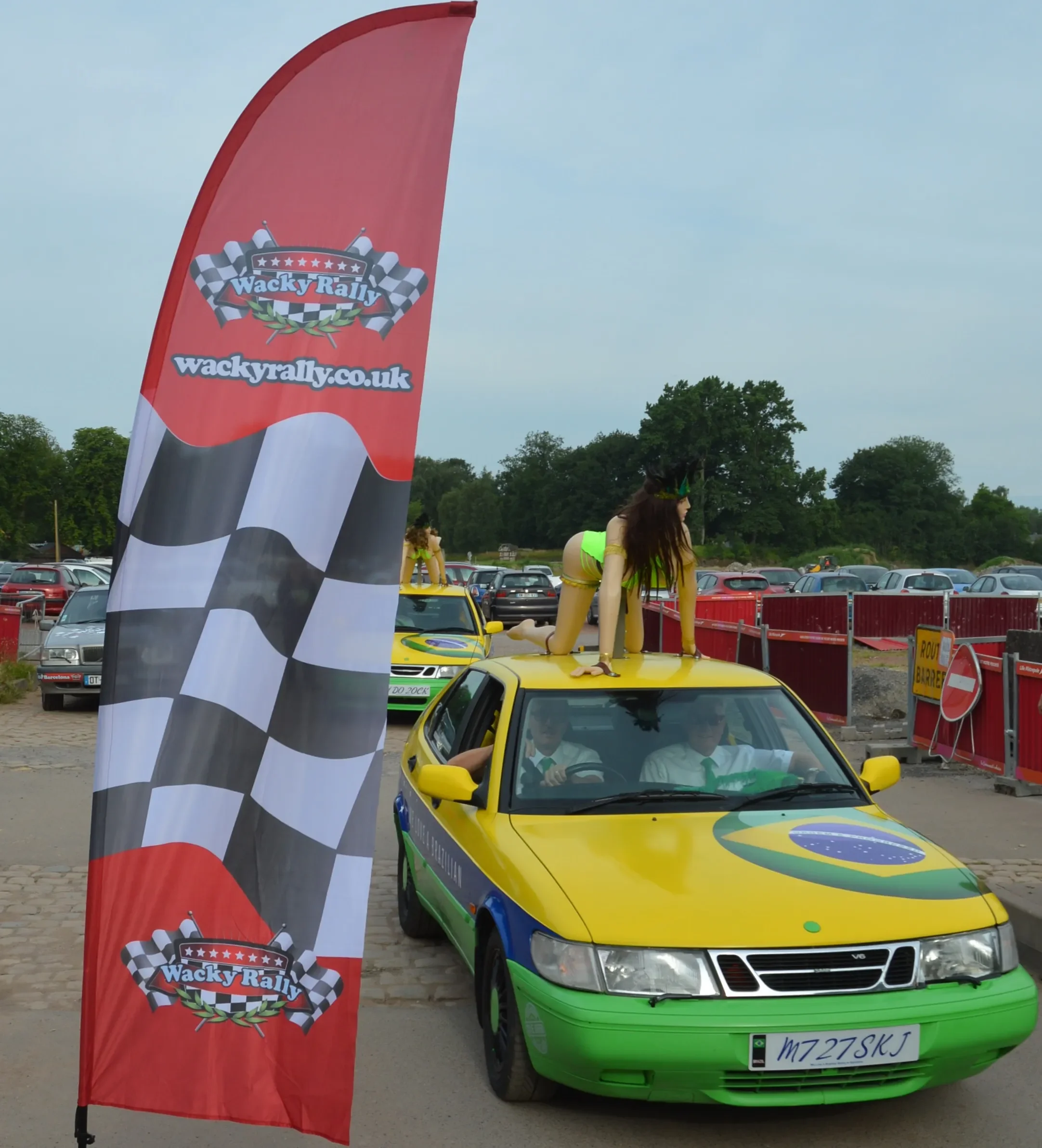A yellow car painted with the Brazilian flag, parked at a rally event with a red Wacky Rally banner in the foreground, and a woman in a revealing costume kneeling on the roof of the car. Several other cars are visible in the background.
