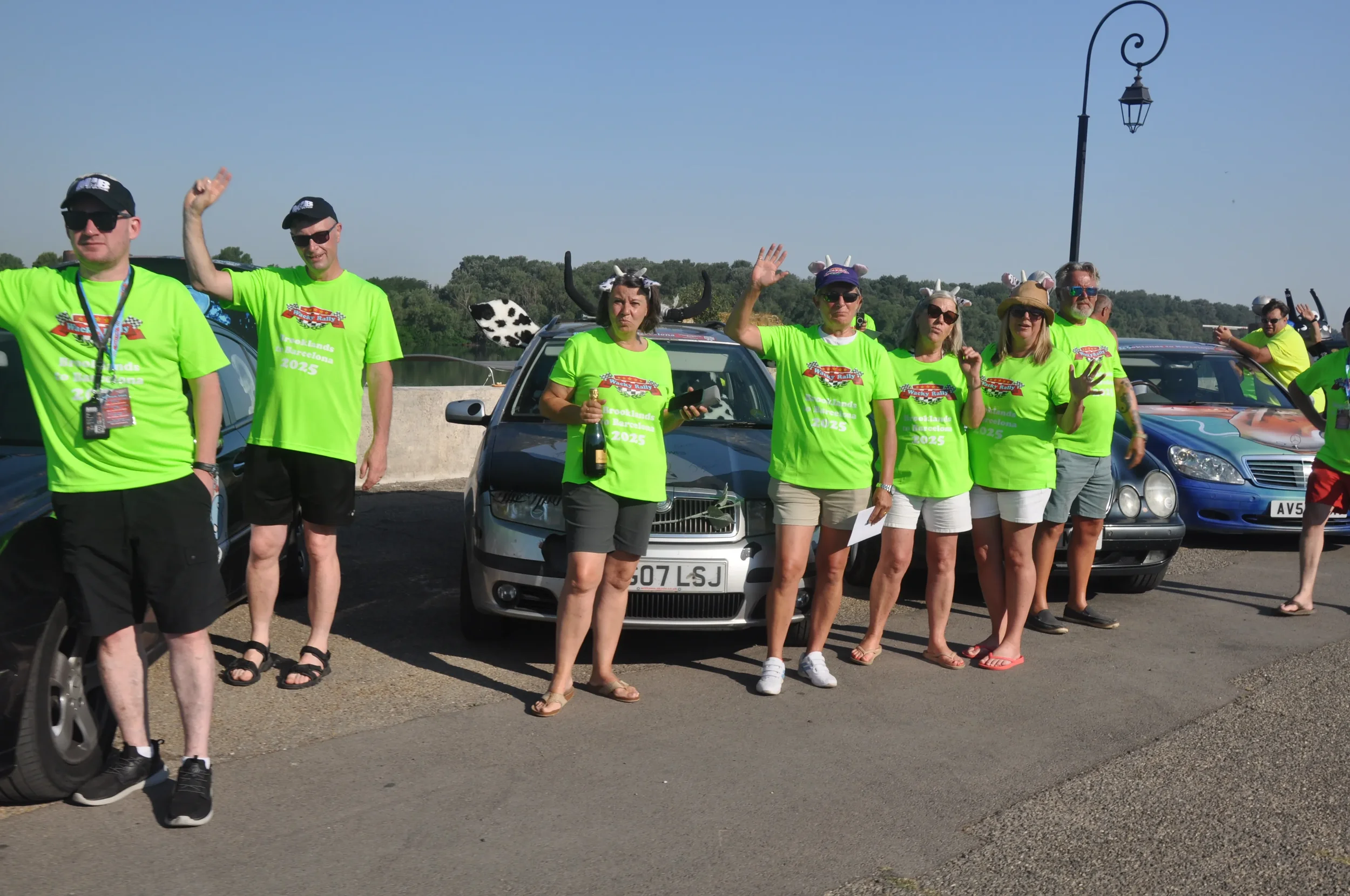 Group of people wearing bright green shirts and animal hats, standing by parked cars on a sunny day to celebrate a car rally event in 2025.