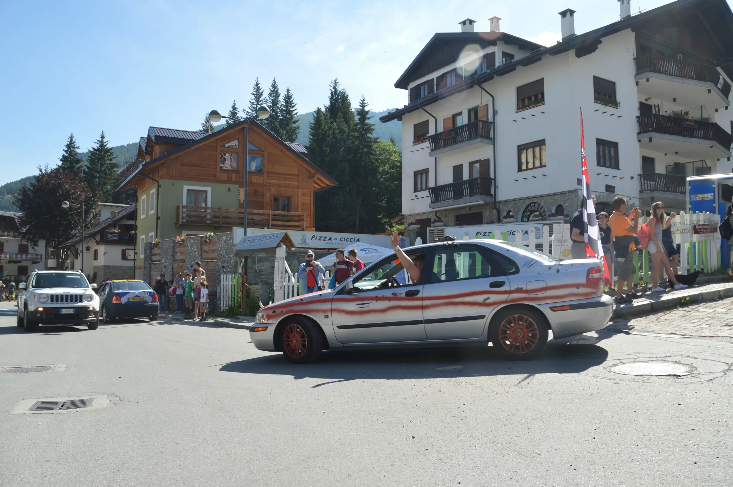 A street scene with a silver car featuring red racing stripes and red wheels, waving a checkered flag, with a crowd of people on the sidewalk and in the background, buildings, and trees.