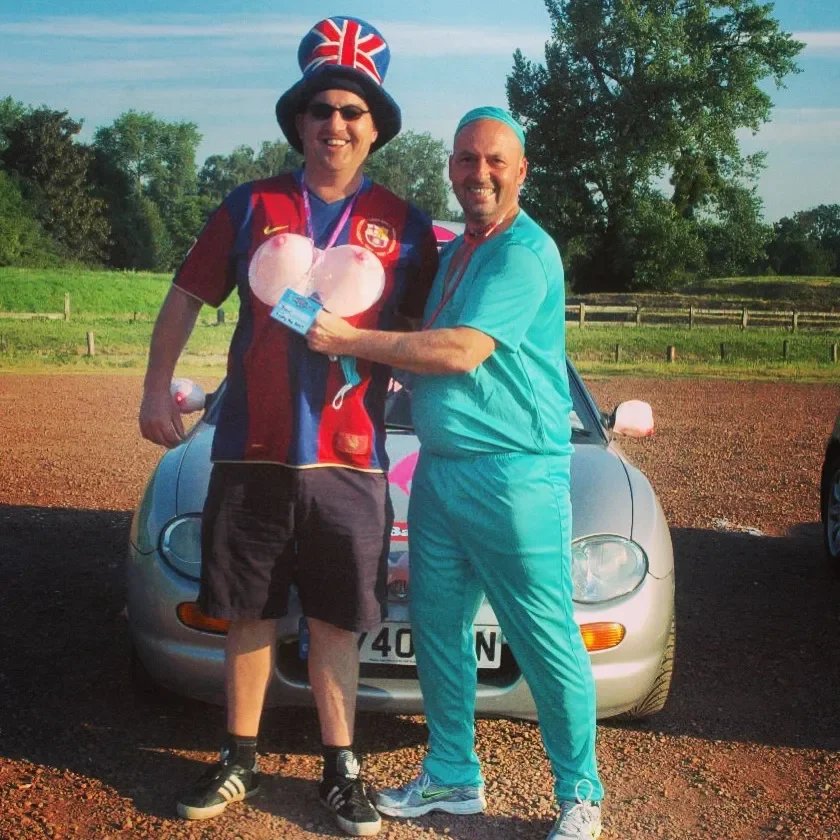 Two men standing in front of a silver car outdoors on a sunny day, smiling and shaking hands. One man is wearing a colorful German football jersey, a large Union Jack-themed hat, sunglasses, and holding a pink balloon, while the other man is dressed 