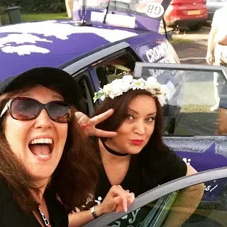 Two women taking a selfie in front of a police car, one making a peace sign and wearing sunglasses, the other wearing a flower crown and looking at the camera.