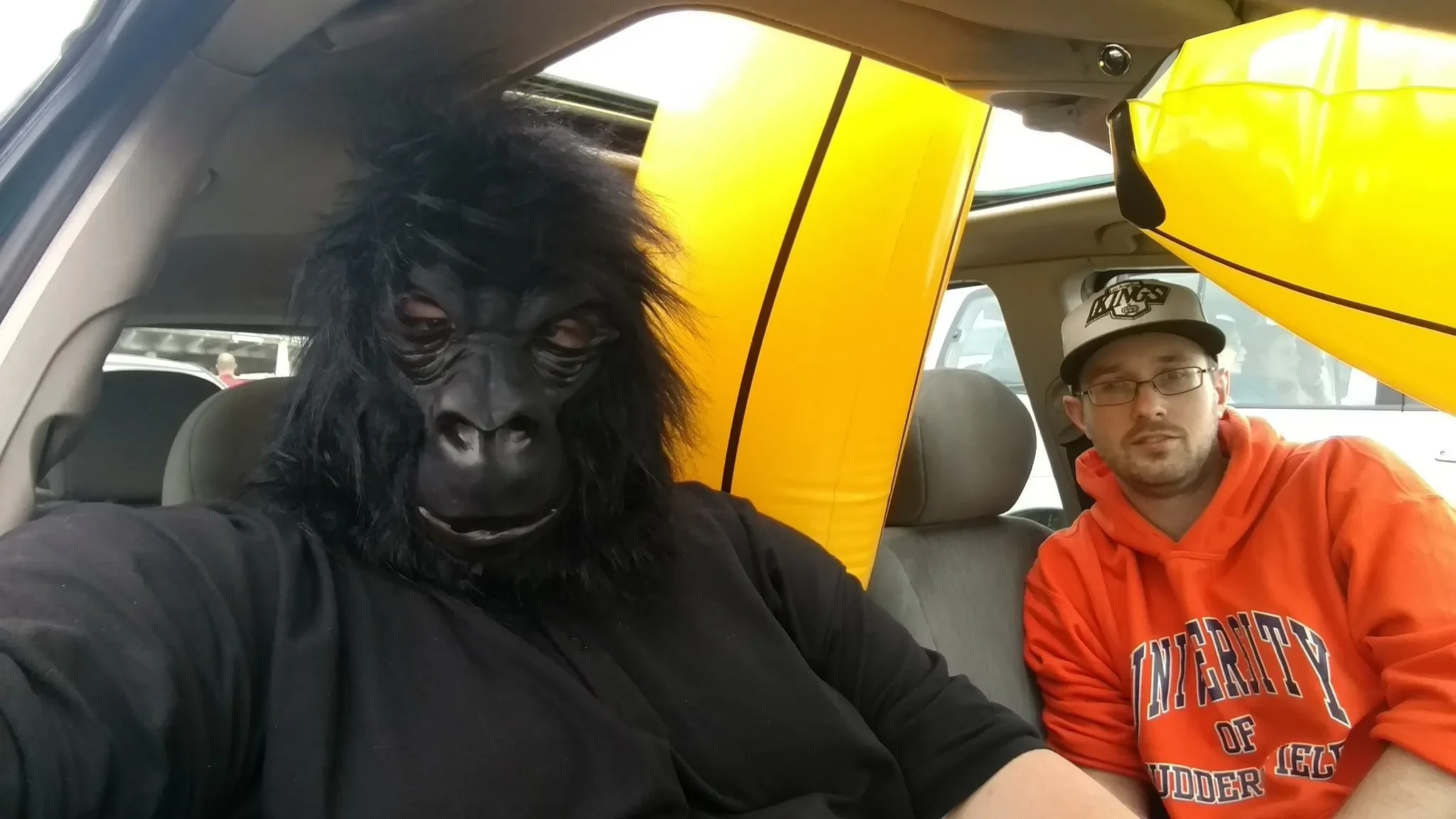 A person in a gorilla mask sitting inside a vehicle, next to a man in an orange hoodie and glasses, with yellow inflatable kayaks in the background.