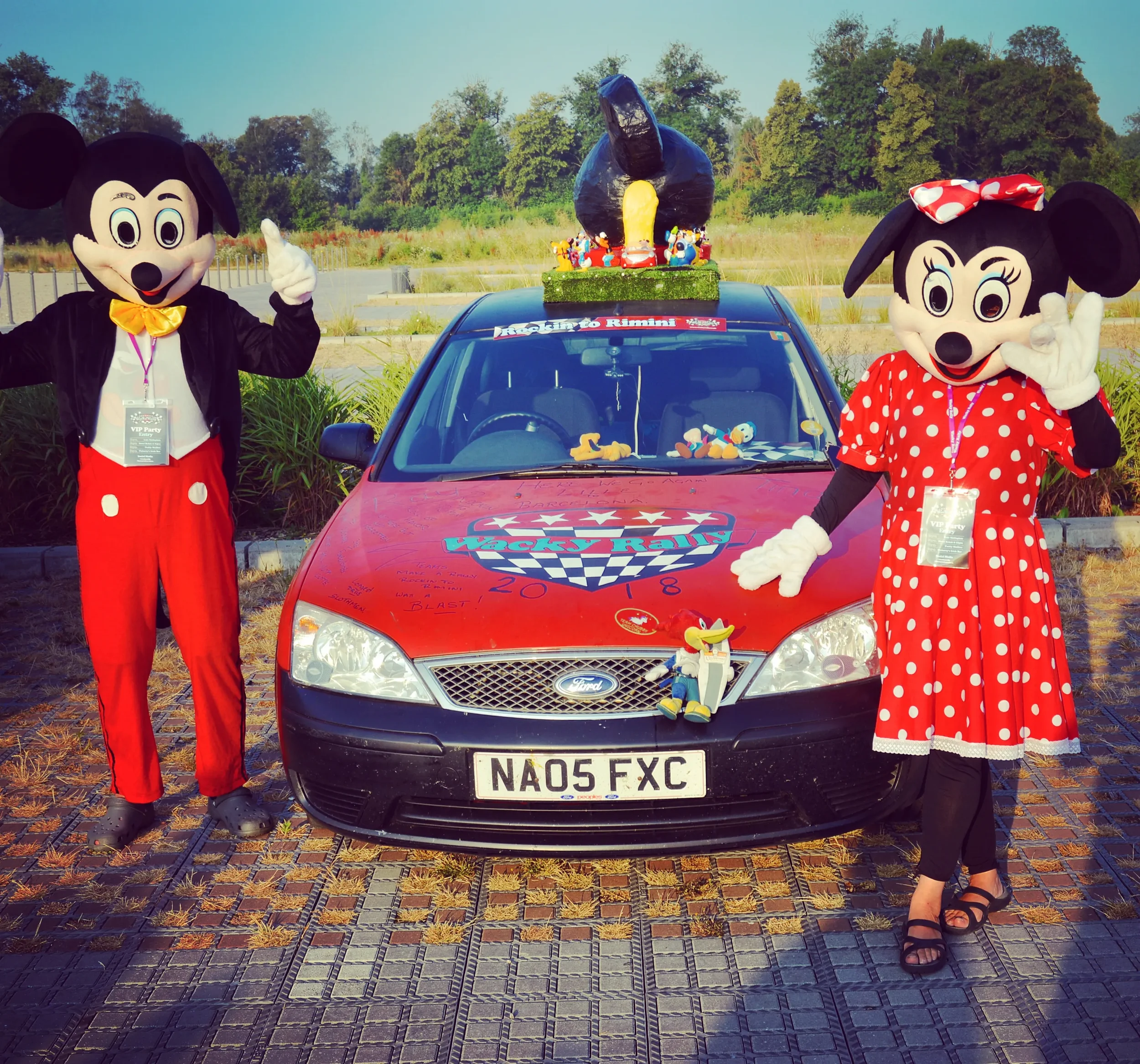 Two people dressed in Mickey and Minnie Mouse costumes posing next to a red Ford car decorated for the Vegas Race event in 2018, with a large Mickey Mouse-themed float on the roof.