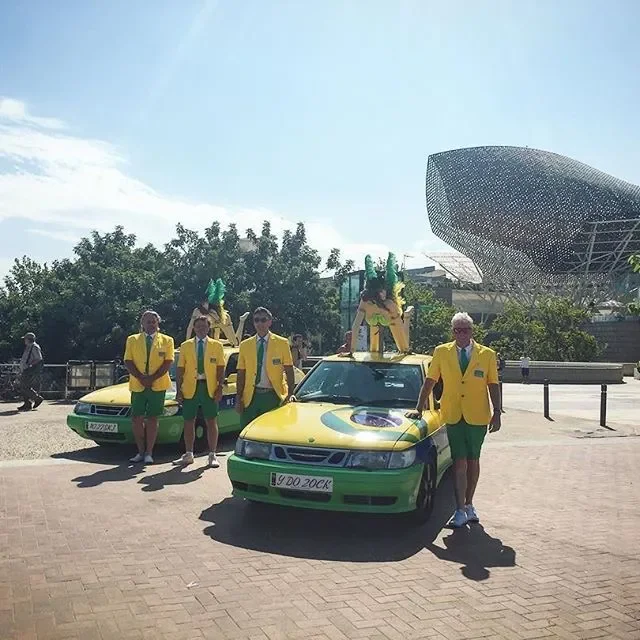 Group of six people dressed in yellow jackets and green shorts, with some wearing green feathered headpieces, standing and sitting on yellow and green cars, in front of a modern architectural building and trees, on a sunny day.