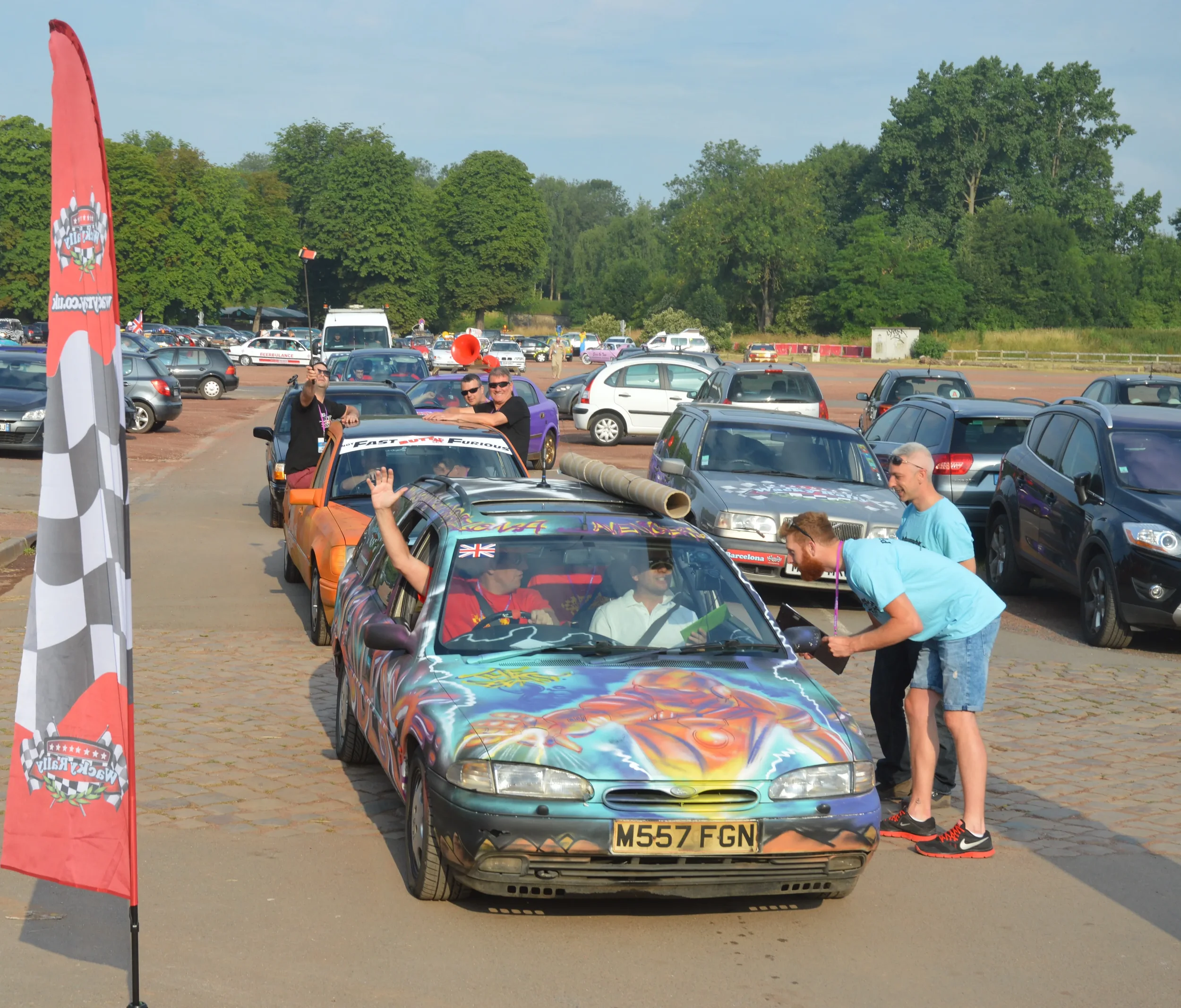 Colorful vintage car with a painted design on the hood and a UK flag decal on the windshield, parked in a line of cars at an outdoor event. People are gathered around the car, some waving and taking photos, with a red and black checkered flag banner 